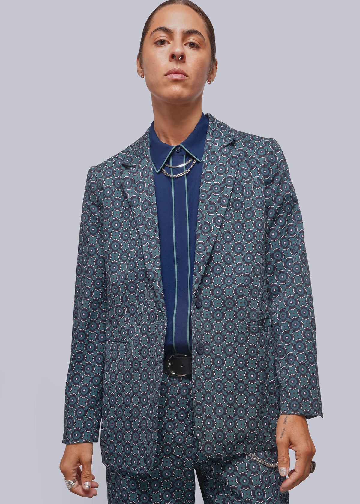 S=A model wearing a tailored blazer and matching trousers in a blue-gray color with a circular medallion pattern, layered over a dark blue shirt and necklace, against a solid gray studio background.