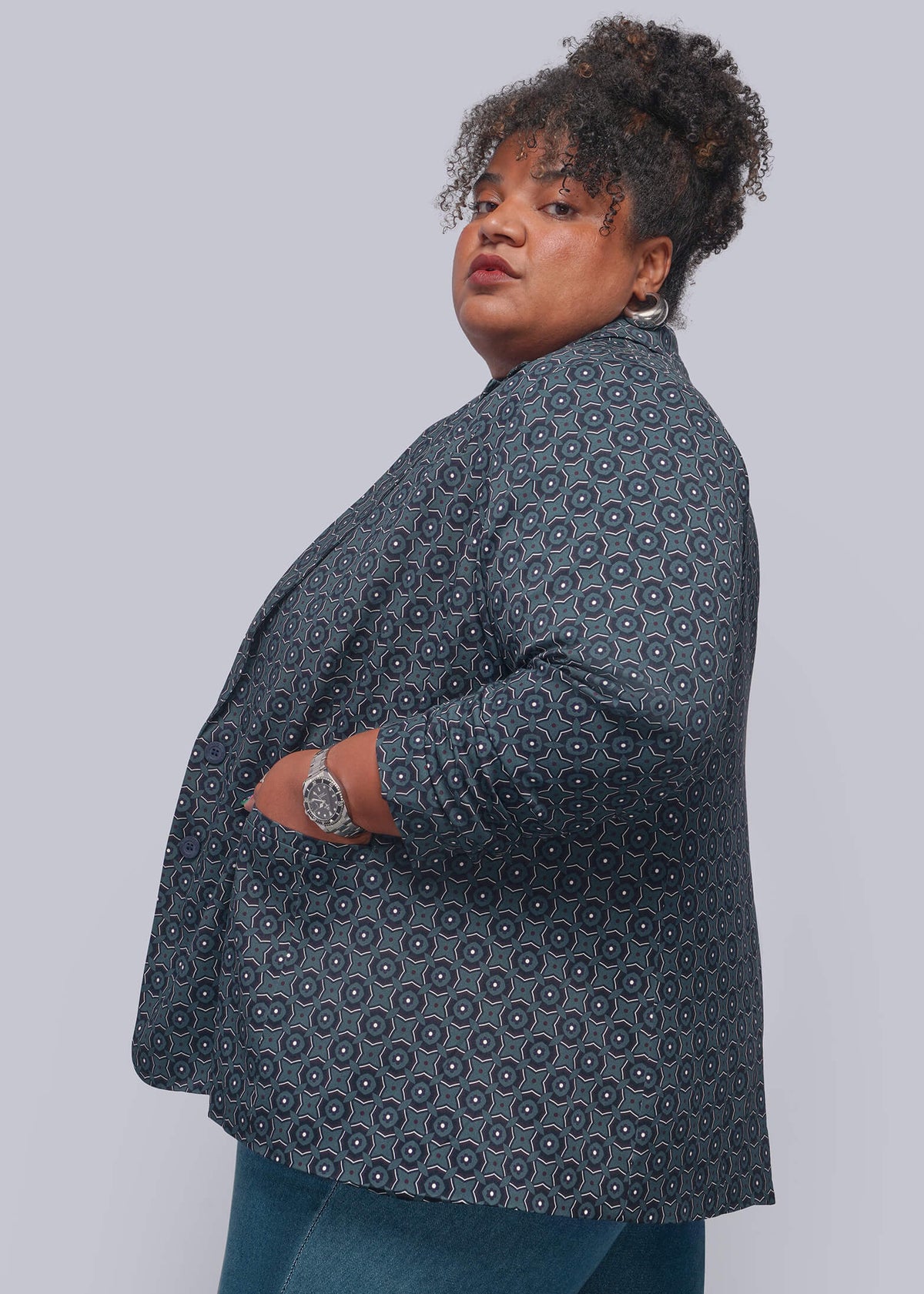 1X=A plus-size model wearing a dark gray patterned blazer and blue jeans, photographed from the side against a solid light gray studio background.
