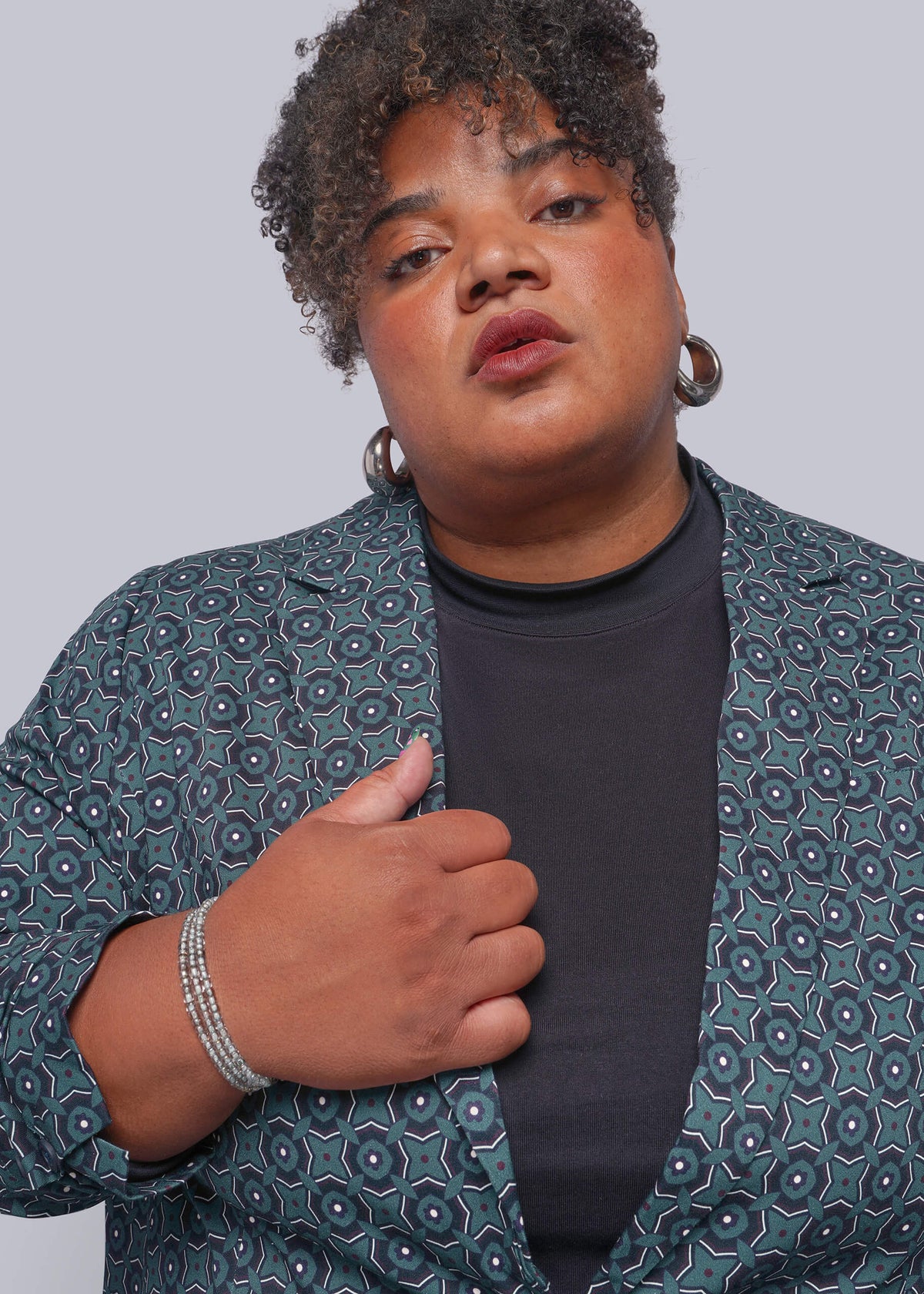 1X=A close-up shot of a person wearing a dark teal patterned blazer over a black crewneck t-shirt, accessorized with silver hoop earrings and a beaded bracelet, against a solid gray background.