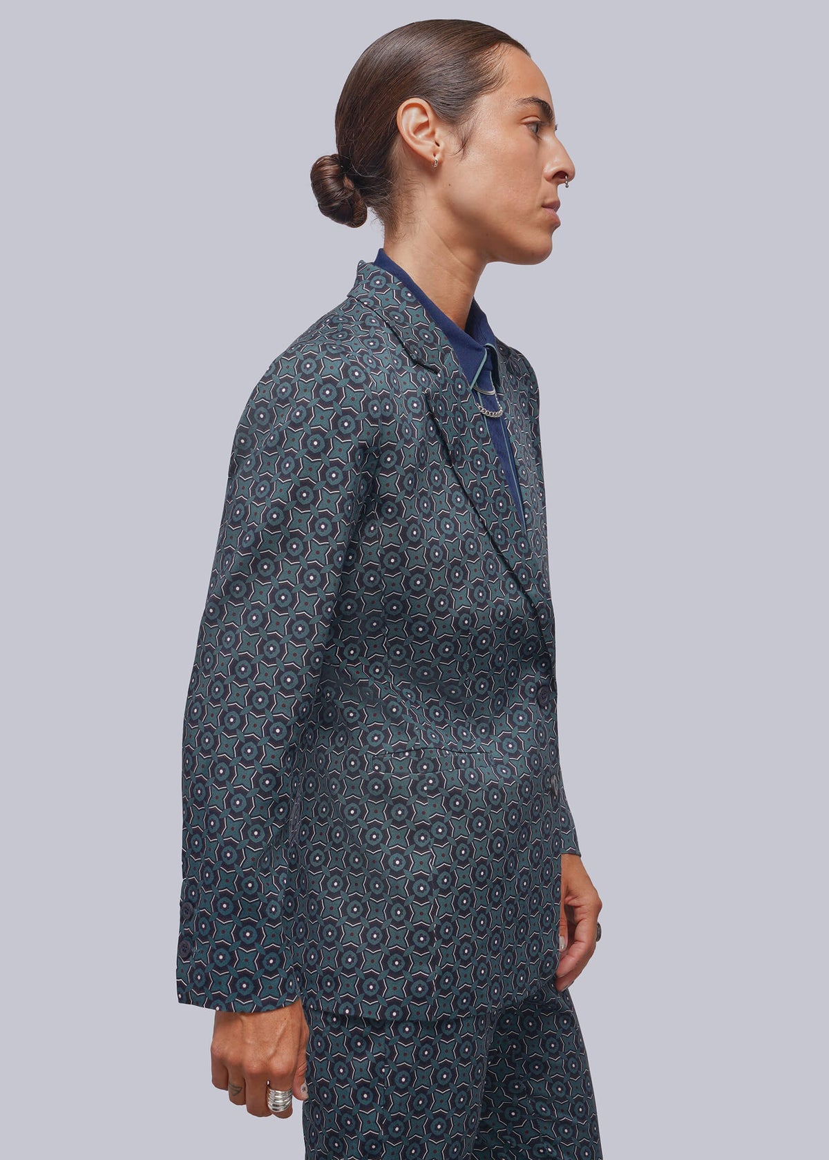 S=A model shown in profile wearing a dark blue patterned suit with a high collar blazer and matching trousers over a navy shirt, set against a solid gray studio background.