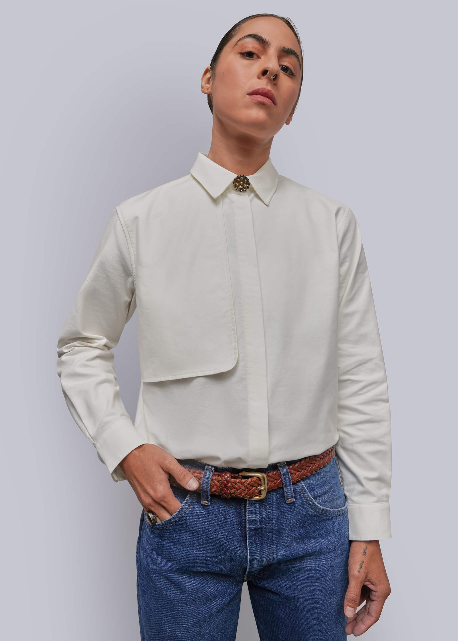 S=A model wearing a white long-sleeve button-down shirt with a chest pocket and a decorative button at the collar, paired with blue jeans and a brown woven belt, against a solid gray background.