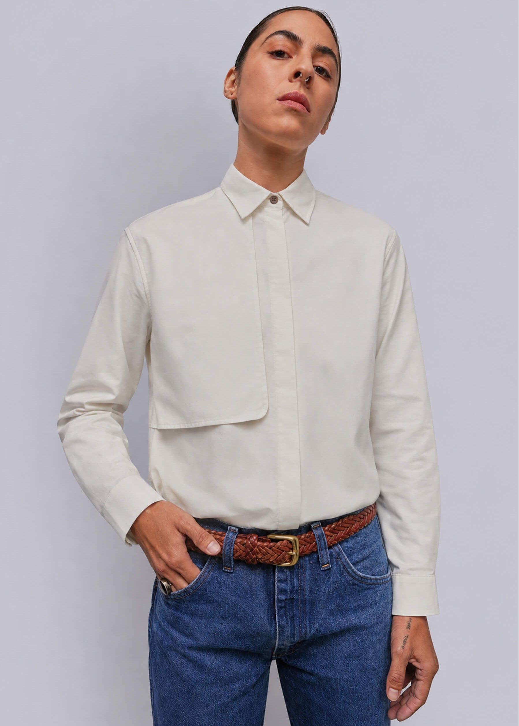 A model wearing a white long-sleeve button-down shirt, paired with blue jeans and a brown woven belt, against a solid gray background.
