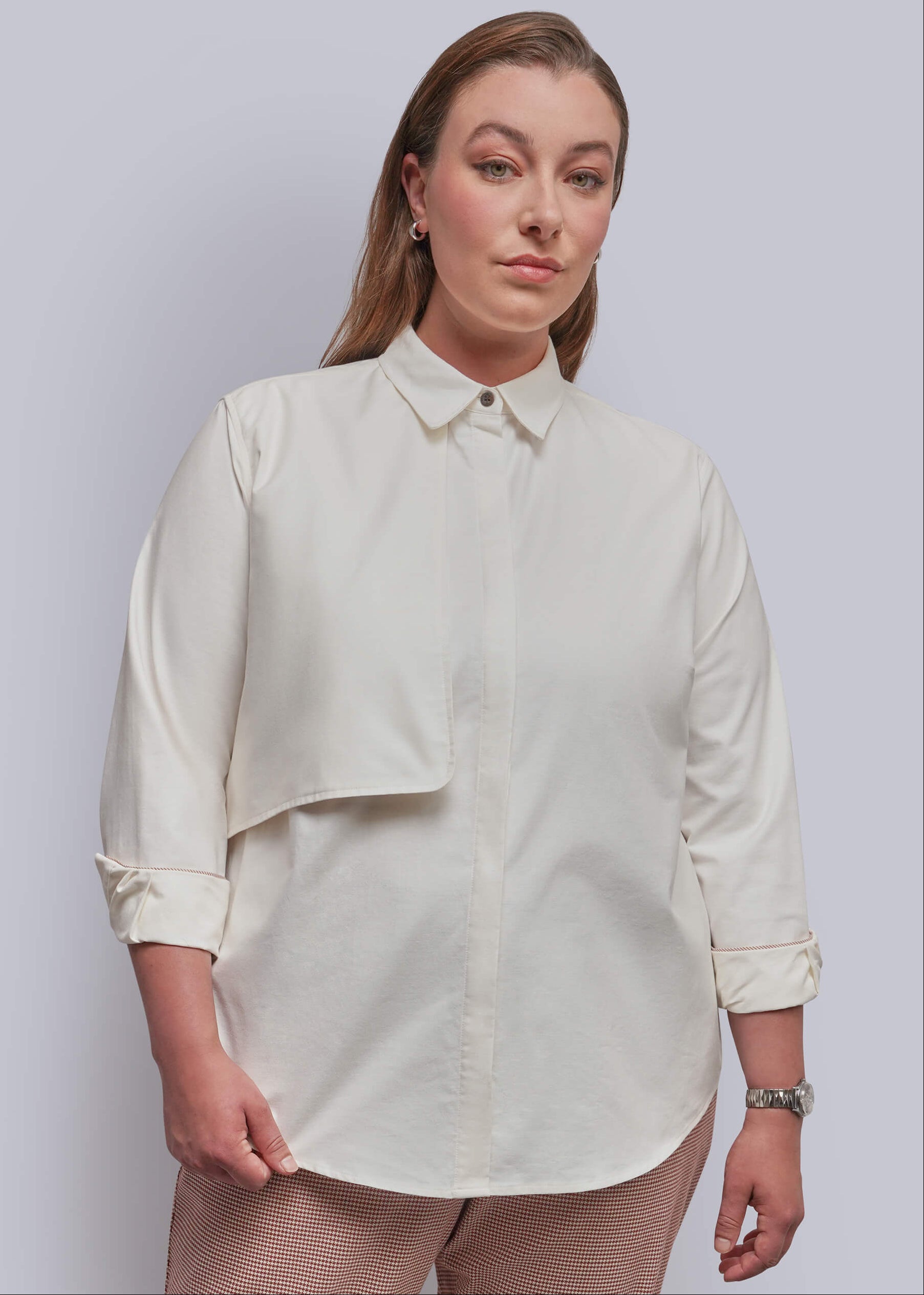 1X=A woman modeling a cream-colored, long-sleeved button-up shirt with a unique asymmetrical flap pocket detail on the chest, paired with beige patterned trousers, against a solid light gray background.