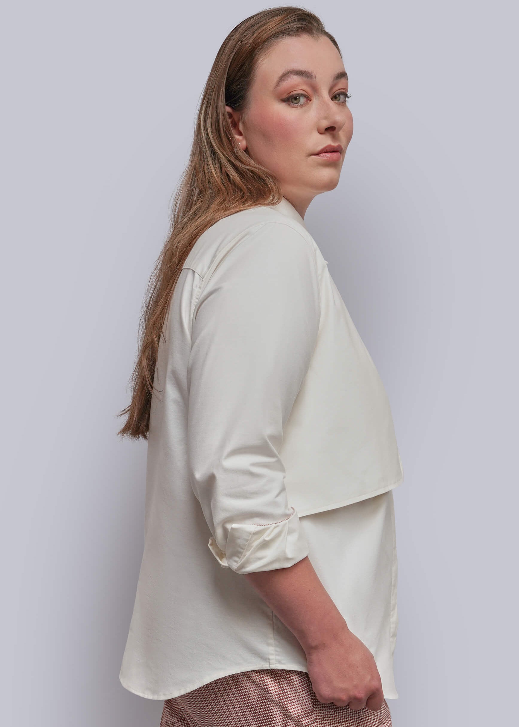 1X=A woman shown from the side and back wearing a white button up, set against a plain light gray studio background.