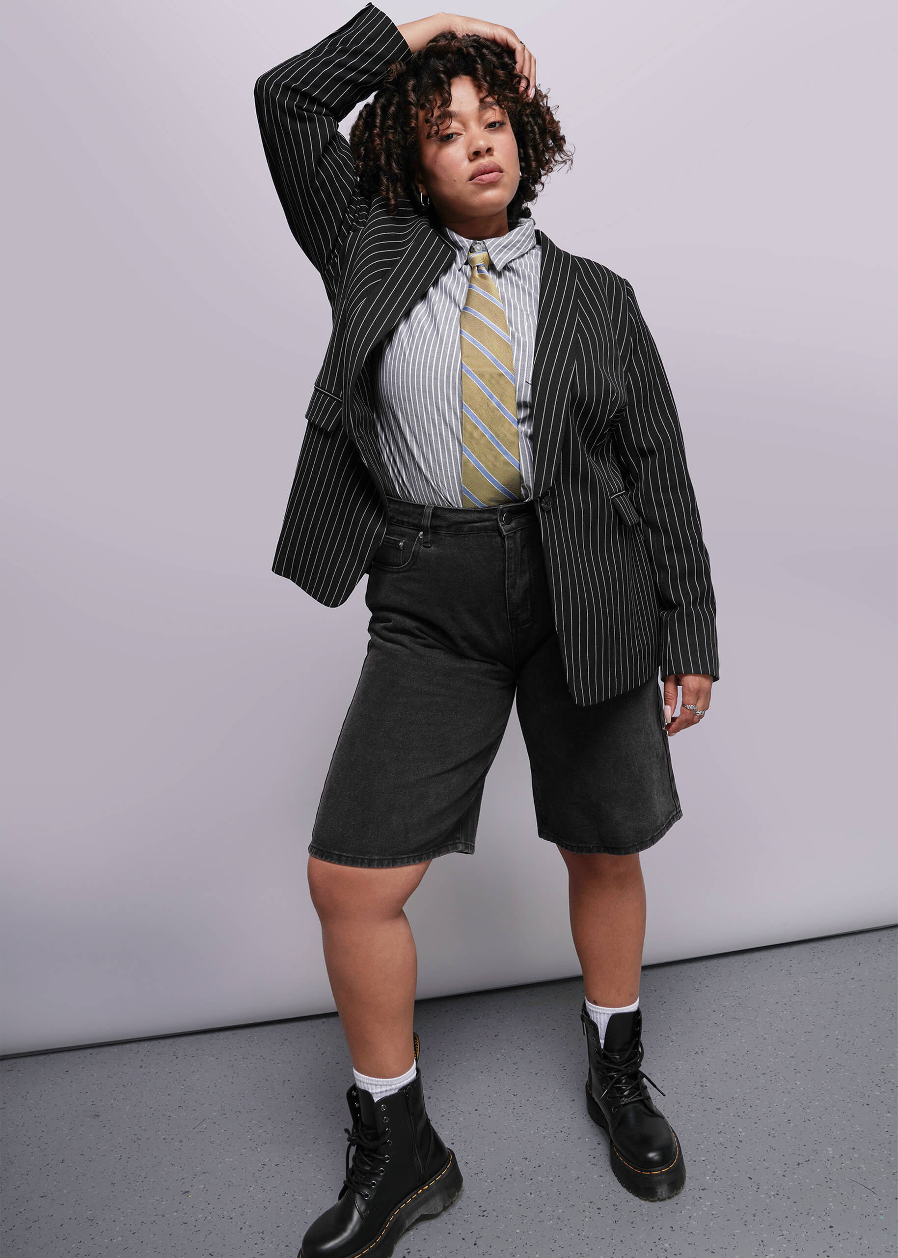 1X=Person wearing a black pinstripe blazer, white shirt with a tie, and black shorts against a plain background