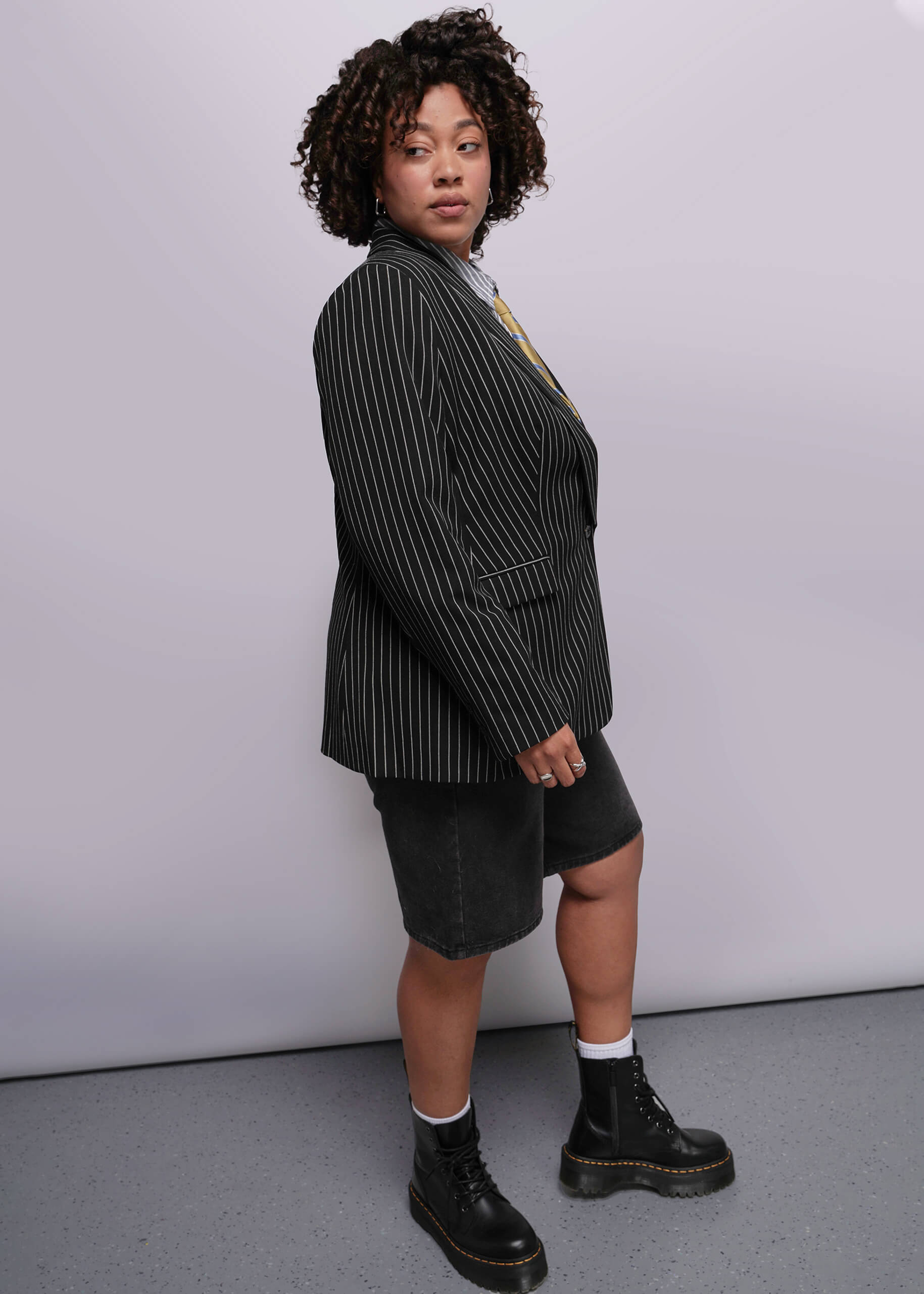 1X=Person wearing a black pinstripe blazer, black shorts, and black boots against a white background