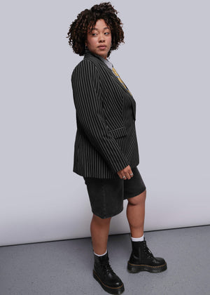 1X=Person wearing a black pinstripe blazer, black shorts, and black boots against a white background