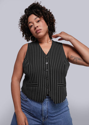 1X=Woman wearing a black pinstripe vest over a blue top against a plain background