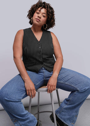 1X=Woman sitting on a stool wearing a black pinstripe vest and blue jeans against a gray background