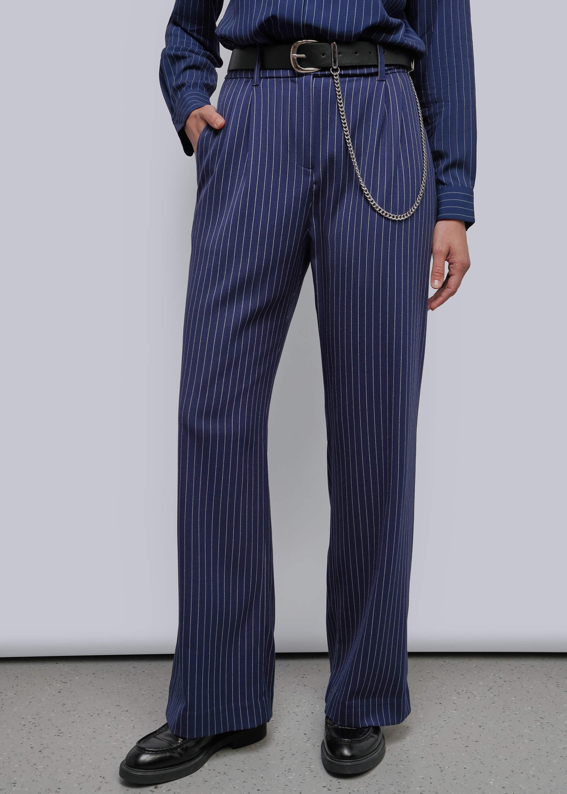 A person wearing navy blue pinstriped trousers with a black belt and silver chain detail, paired with a matching navy shirt and black shoes, photographed against a plain light gray studio background.