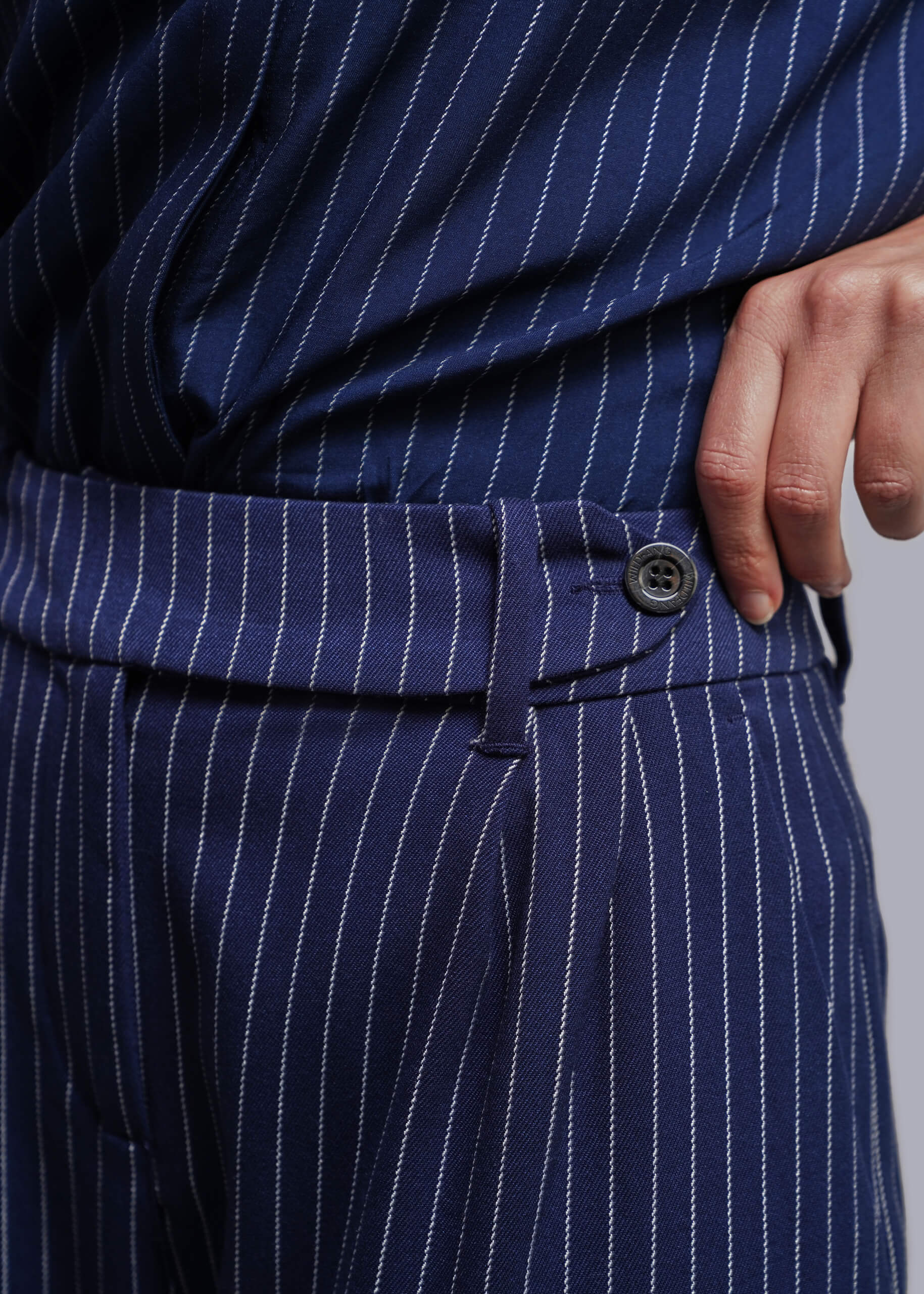 S=Close-up of navy blue pinstriped trousers with a matching top, showing the waistband, button, and a hand resting near the pocket.