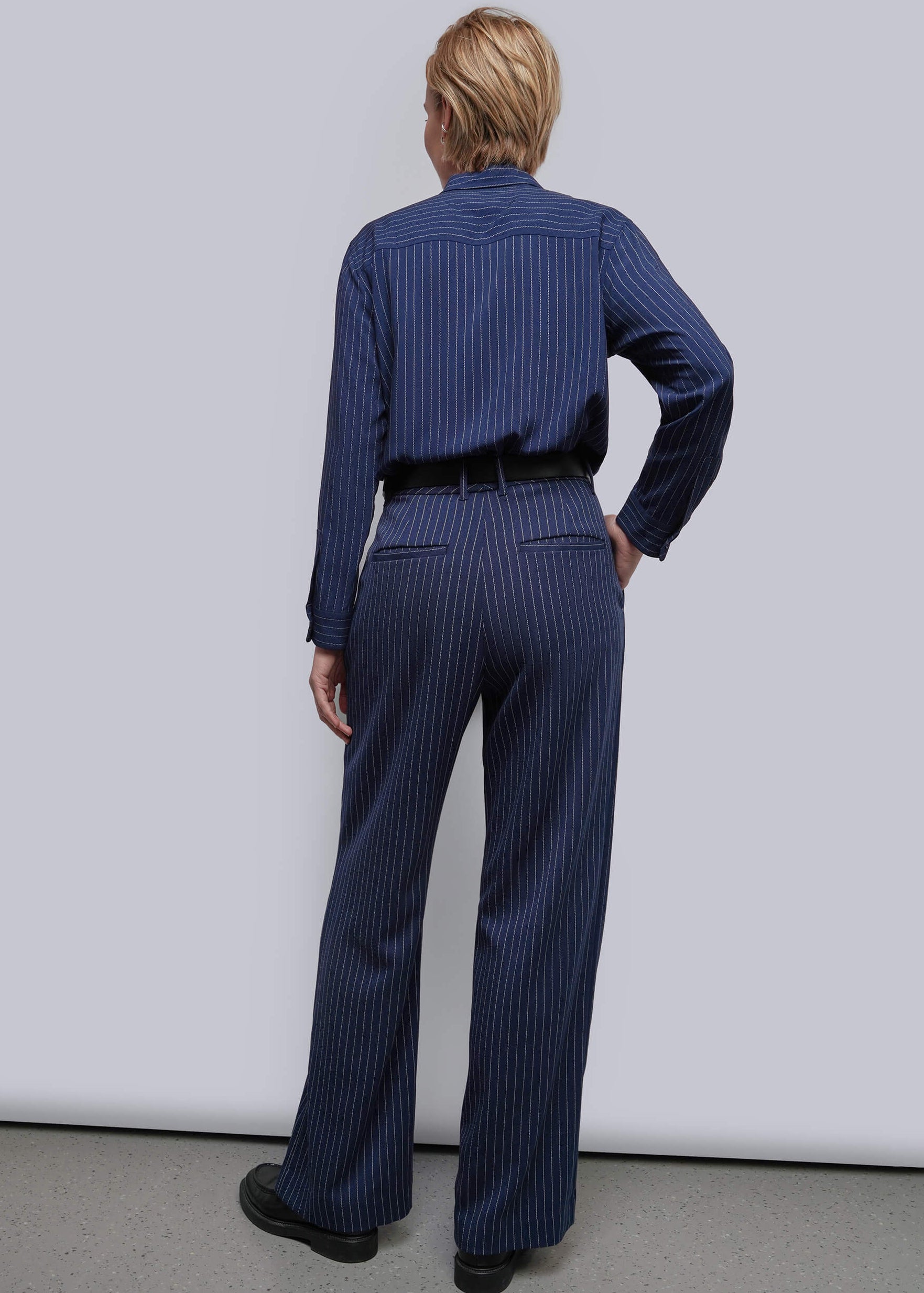 S=A model viewed from the back wearing a navy blue pinstripe long-sleeve shirt and matching pinstripe trousers, paired with a black belt and black shoes, against a light gray studio background.