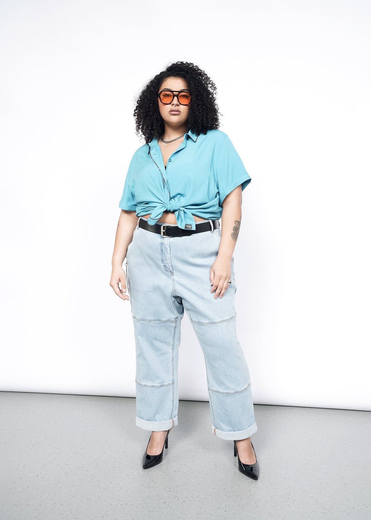 Model wearing the Empower Button Up in Coastal with Floral print piped trim in size 1X, tied in a knot at waist, with jeans, a black belt, black pumps, and sunglasses