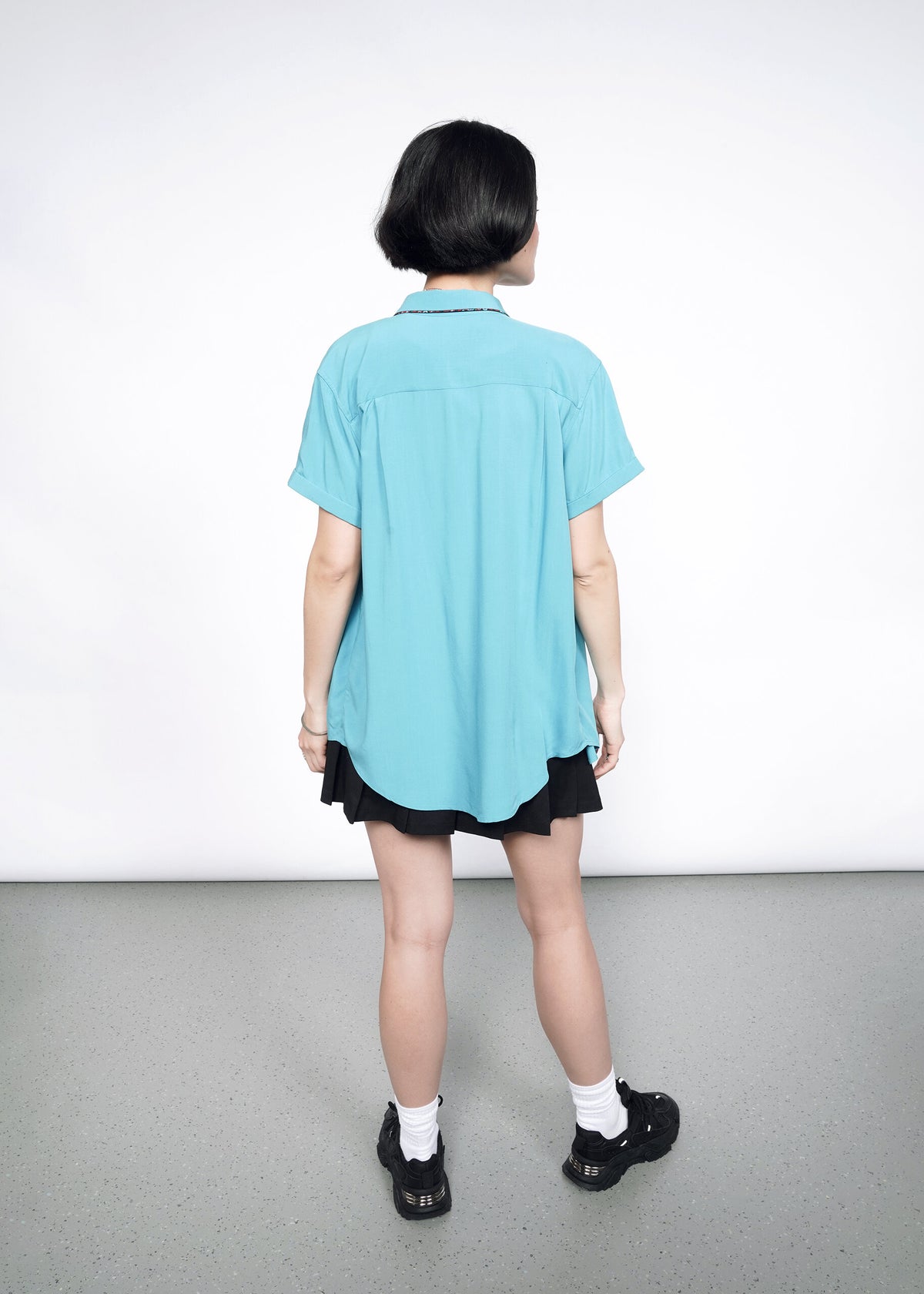 Back image of Model wearing the Empower Button Up in Coastal with Floral print piped trim in size XS, with a black skirt, white socks, and black shoes.