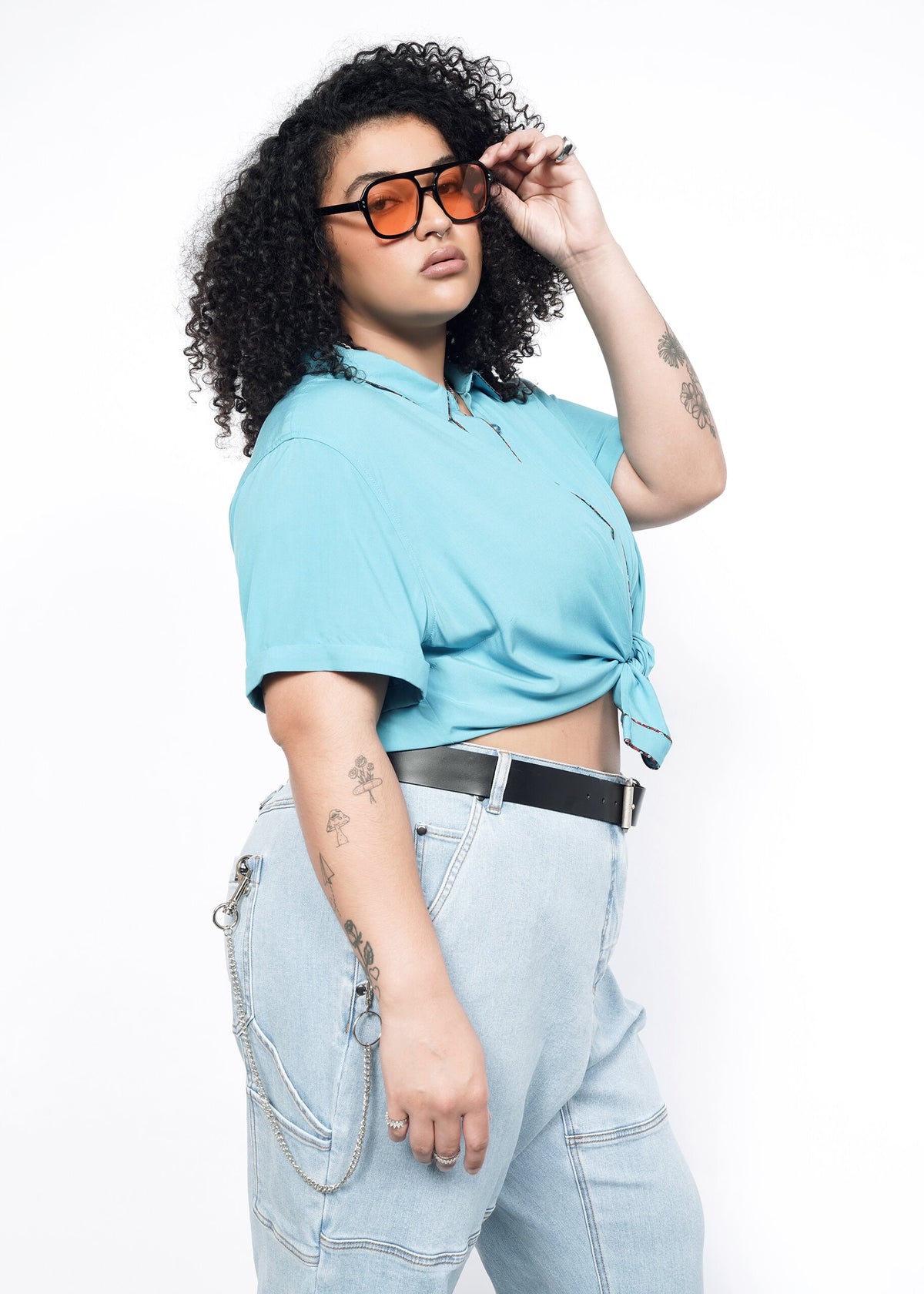 Side image of Model wearing the Empower Button Up in Coastal with Floral print piped trim in size 1X, tied in a knot at waist, with jeans, a black belt, and sunglasses