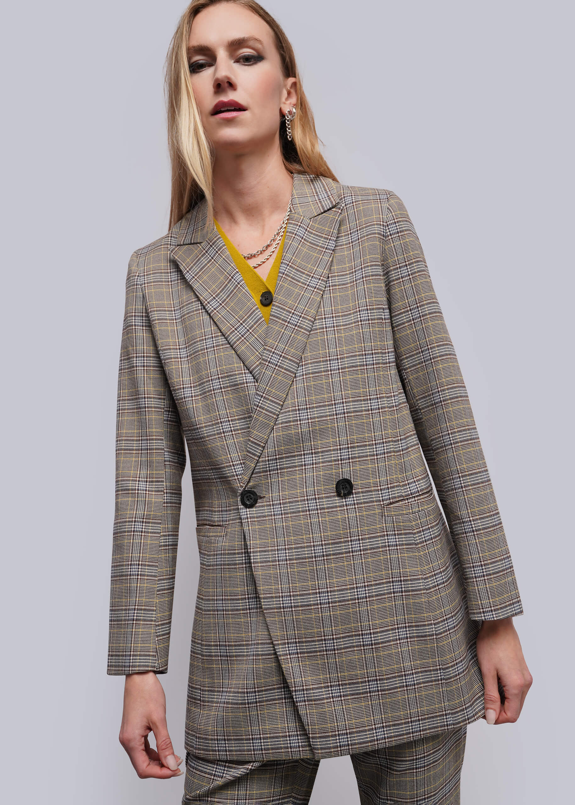 A woman wearing a double-breasted plaid blazer in beige, brown, and gray tones, styled with a yellow top and layered necklaces, against a solid gray studio background.