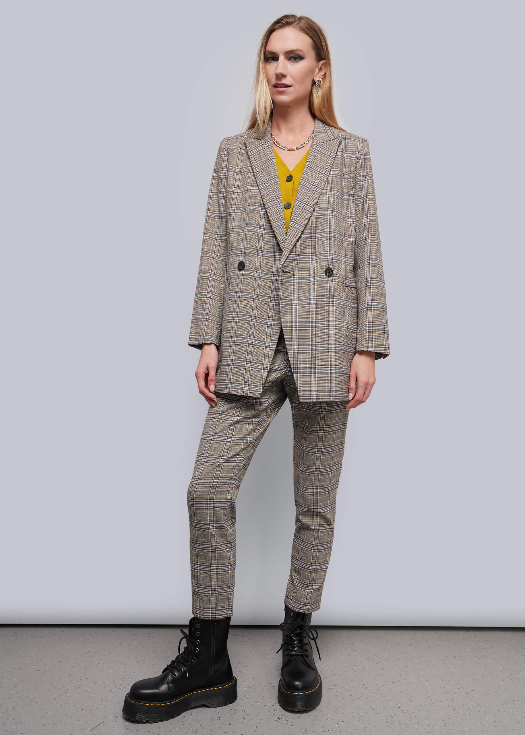 A woman modeling a beige and brown plaid double-breasted blazer with matching trousers, worn over a yellow top and paired with black combat boots, against a plain gray studio background.