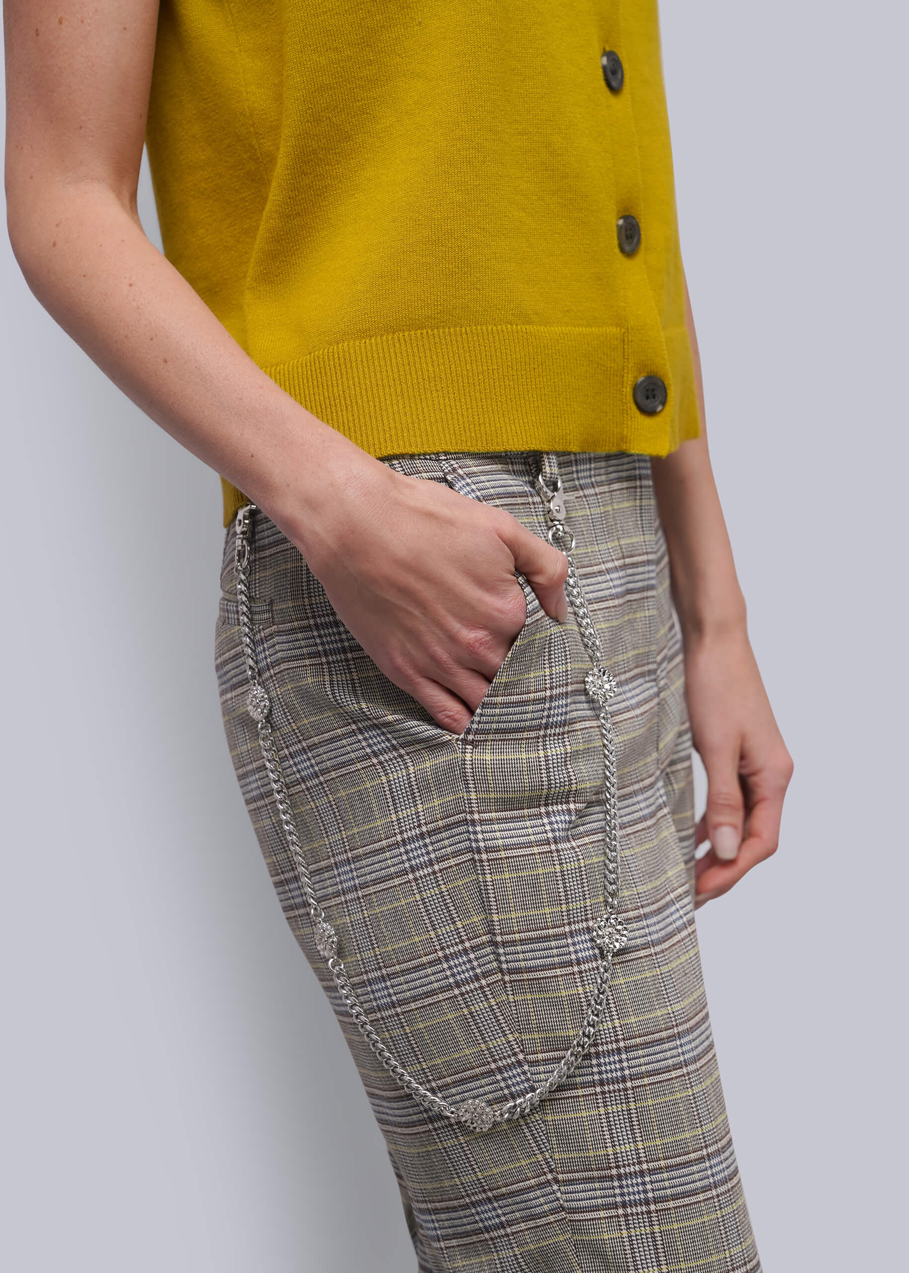 S=A close-up of a person wearing a mustard yellow sleeveless cardigan and beige plaid pants with silver chain details, against a light gray background.