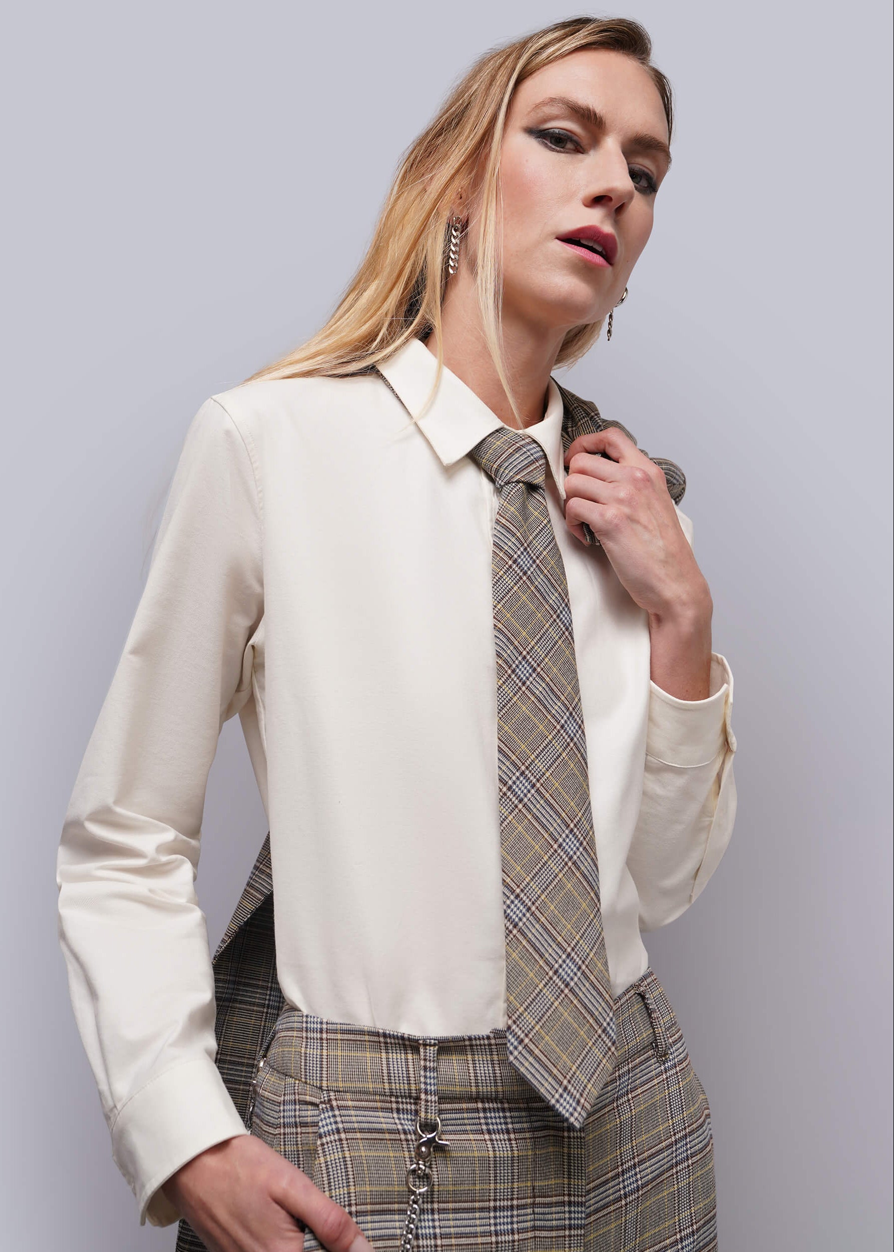 A woman modeling a cream long-sleeve blouse paired with a brown and beige plaid tie and matching plaid trousers, set against a solid light gray studio background.