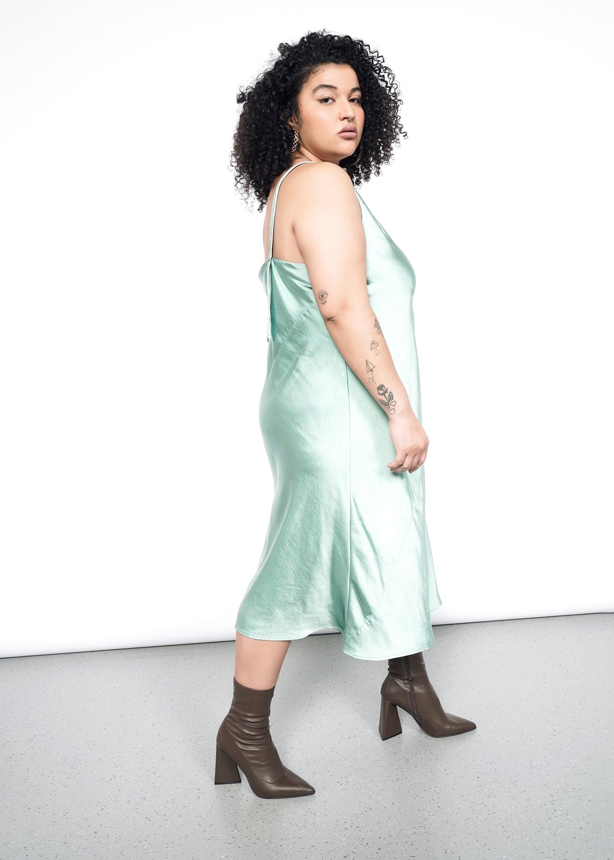 Side image of Model wearing the Empower Satin Slip Dress in Sage in size 1X with heeled ankle boots.