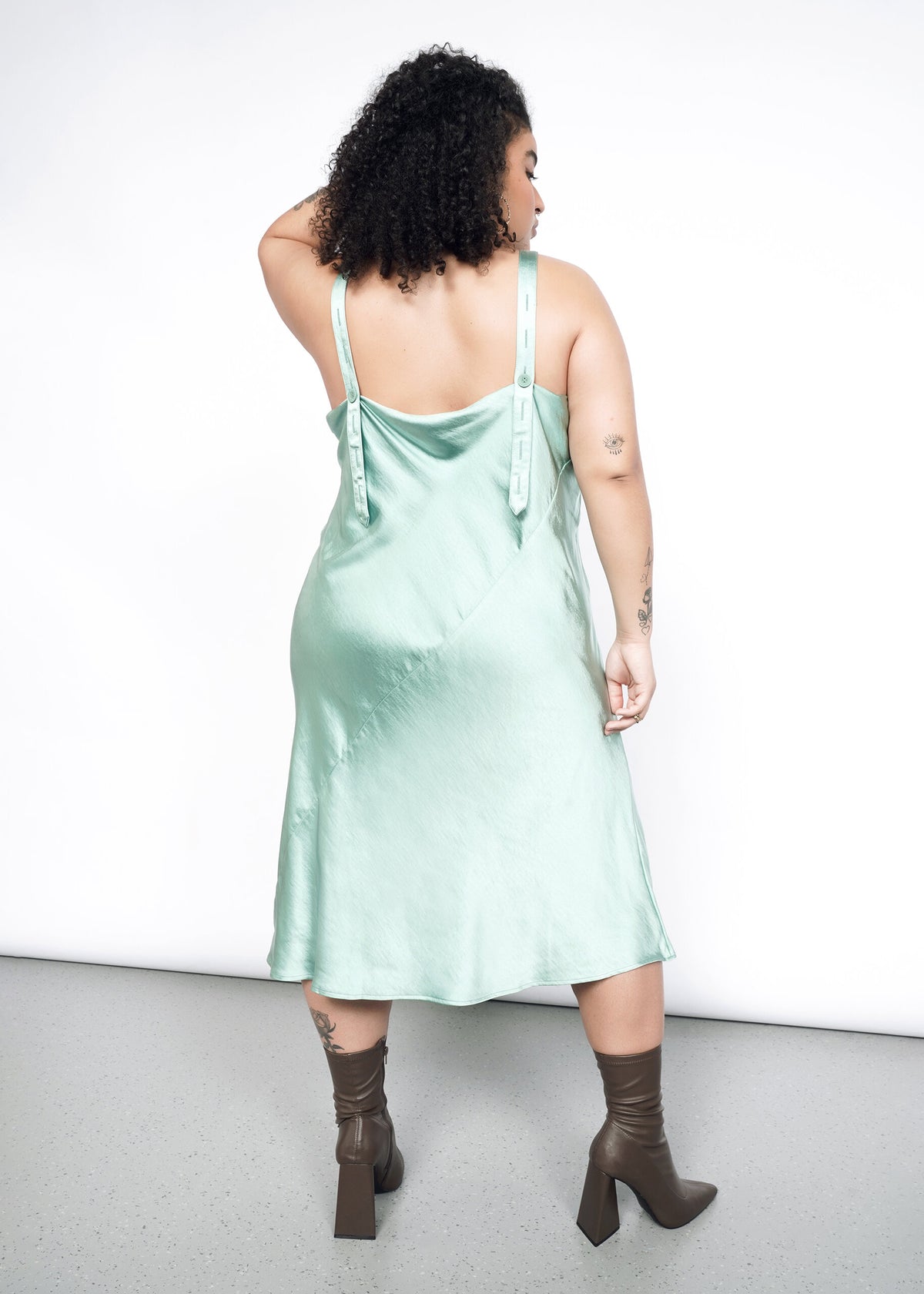 Back image of Model wearing the Empower Satin Slip Dress in Sage in size 1X with heeled ankle boots.