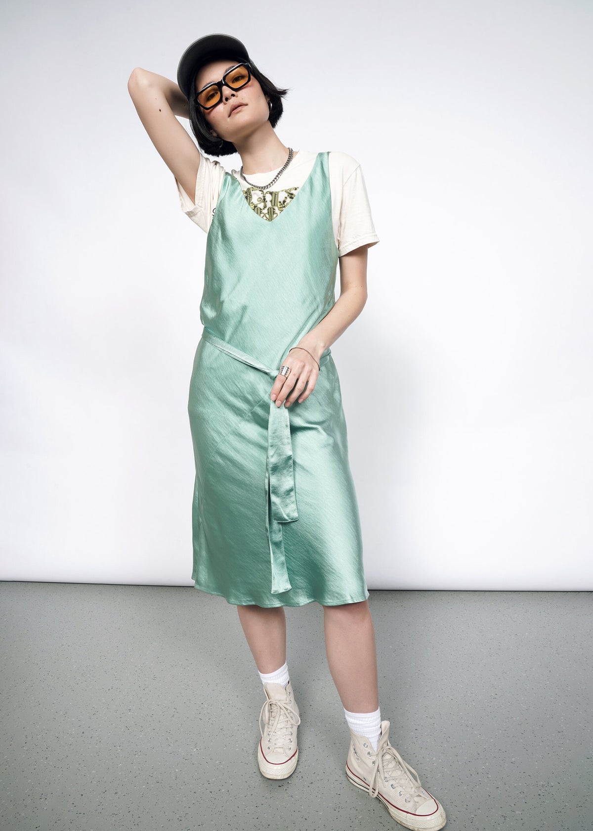 Model wearing the Empower Satin Slip Dress in Sage in size S with a white graphic T, white socks and sneakers, sunglasses, and a ballcap.