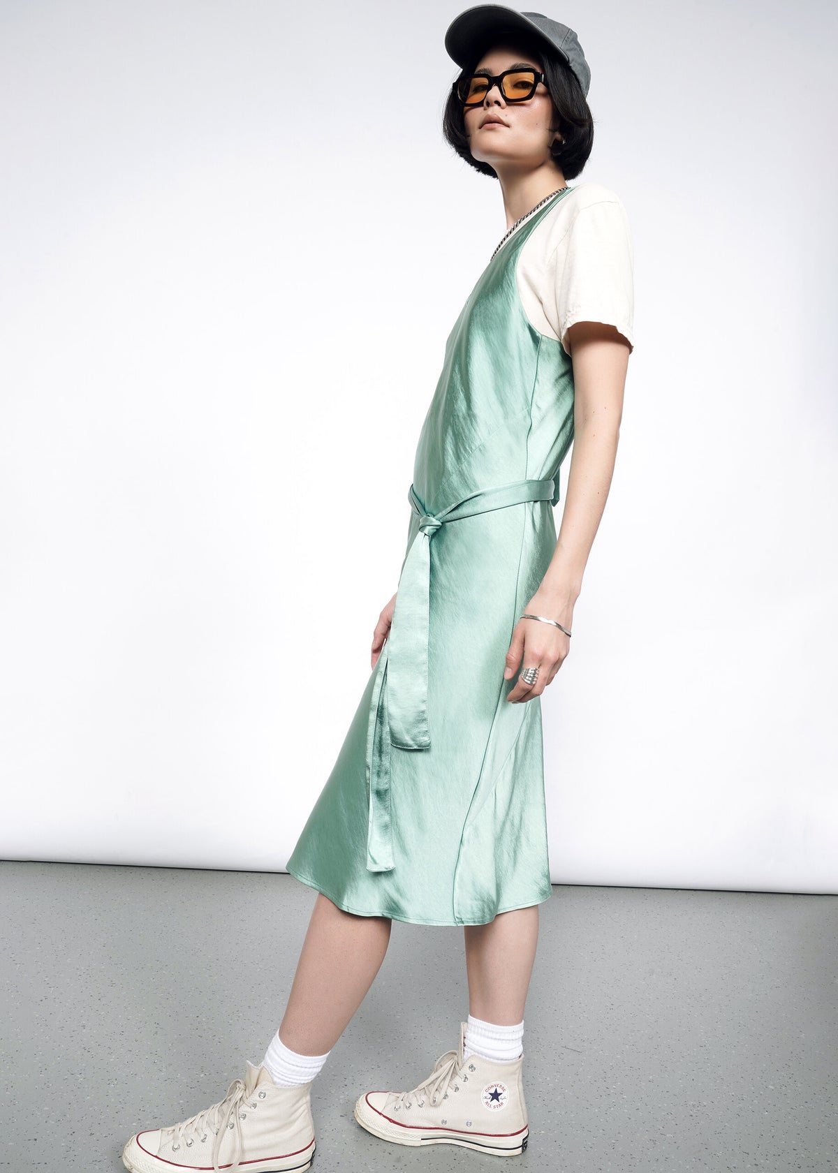Side image of Model wearing the Empower Satin Slip Dress in Sage in size S with a white graphic T, white socks and sneakers, sunglasses, and a ballcap.