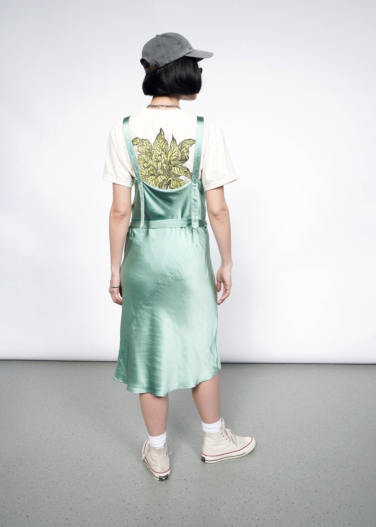 Back image of Model wearing the Empower Satin Slip Dress in Sage in size S with a white graphic T, white socks and sneakers, sunglasses, and a ballcap.