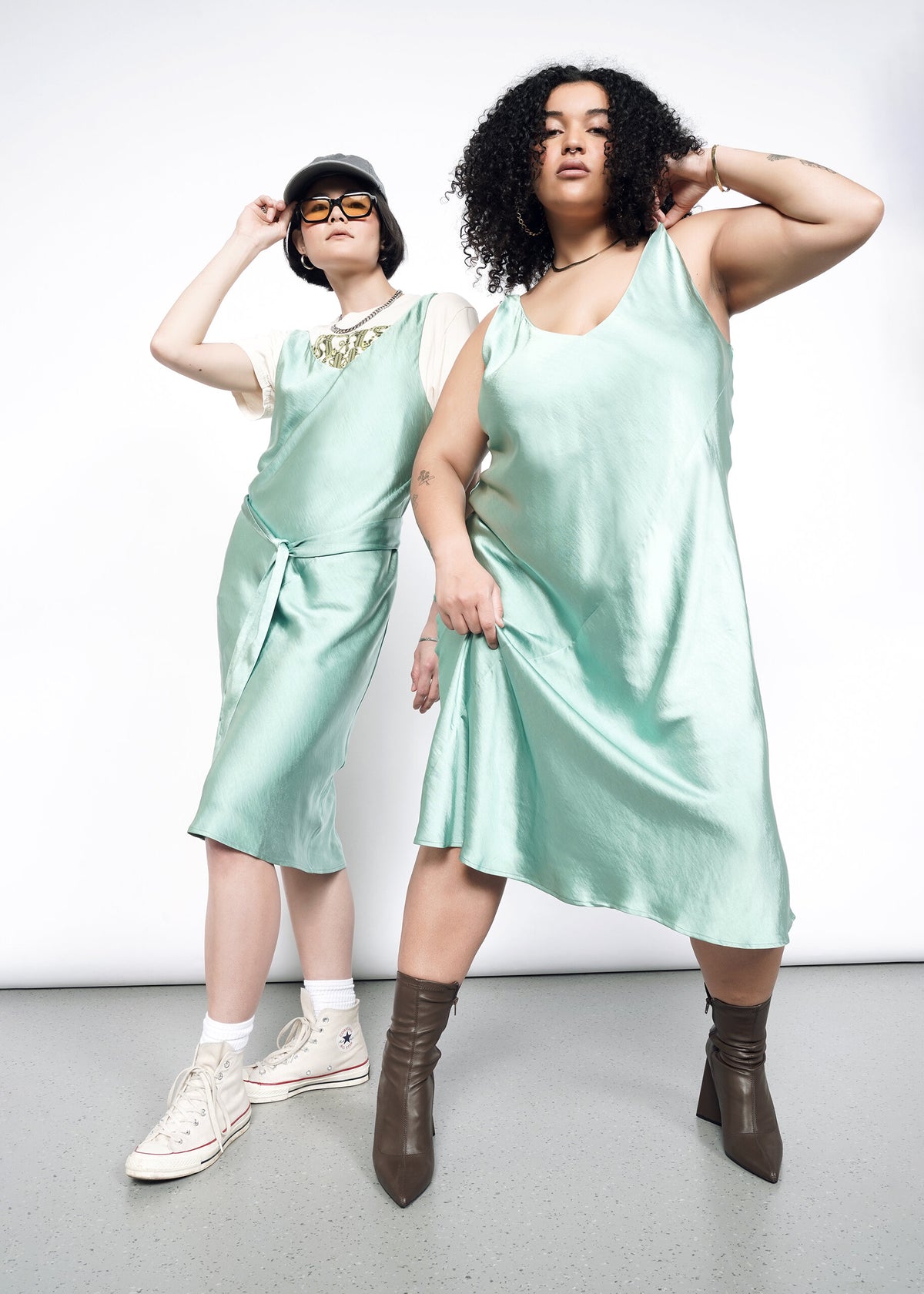 2 Models wearing the Empower Satin Slip Dress in Sage. One in size 1X with heeled ankle boots. One in size S with a white graphic T, white socks and sneakers, sunglasses, and a ballcap. 