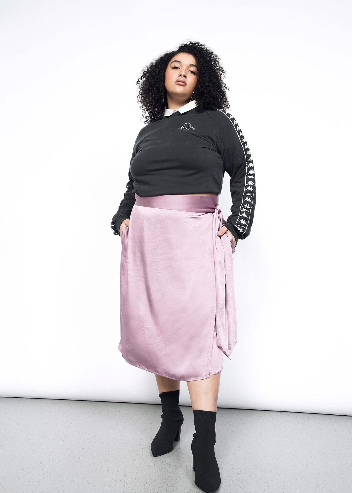 Model wearing the Empower Satin Wrap Skirt in Mauve in size 1X, with a black and white sweatshirt, and black heeled ankle boots.