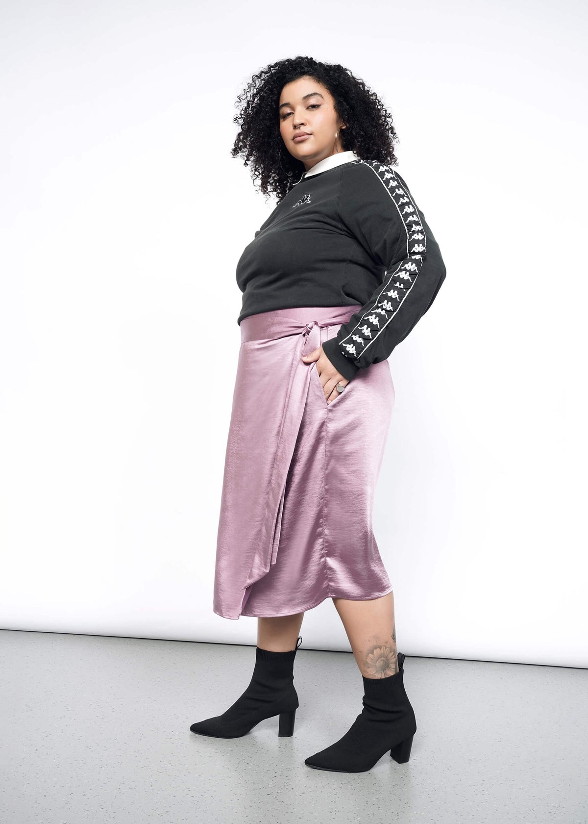Side image of Model wearing the Empower Satin Wrap Skirt in Mauve in size 1X, with a black and white sweatshirt, and black heeled ankle boots.