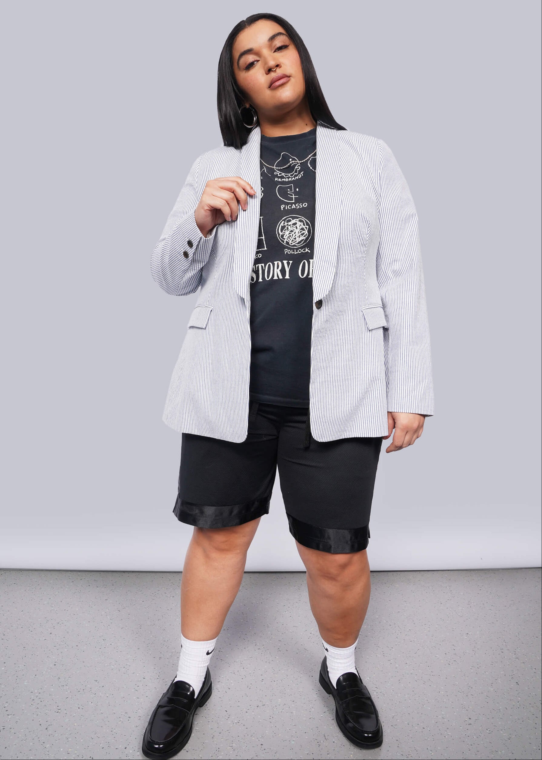 1X=Person wearing a light blue striped blazer over a black graphic t-shirt with black shorts on a plain background