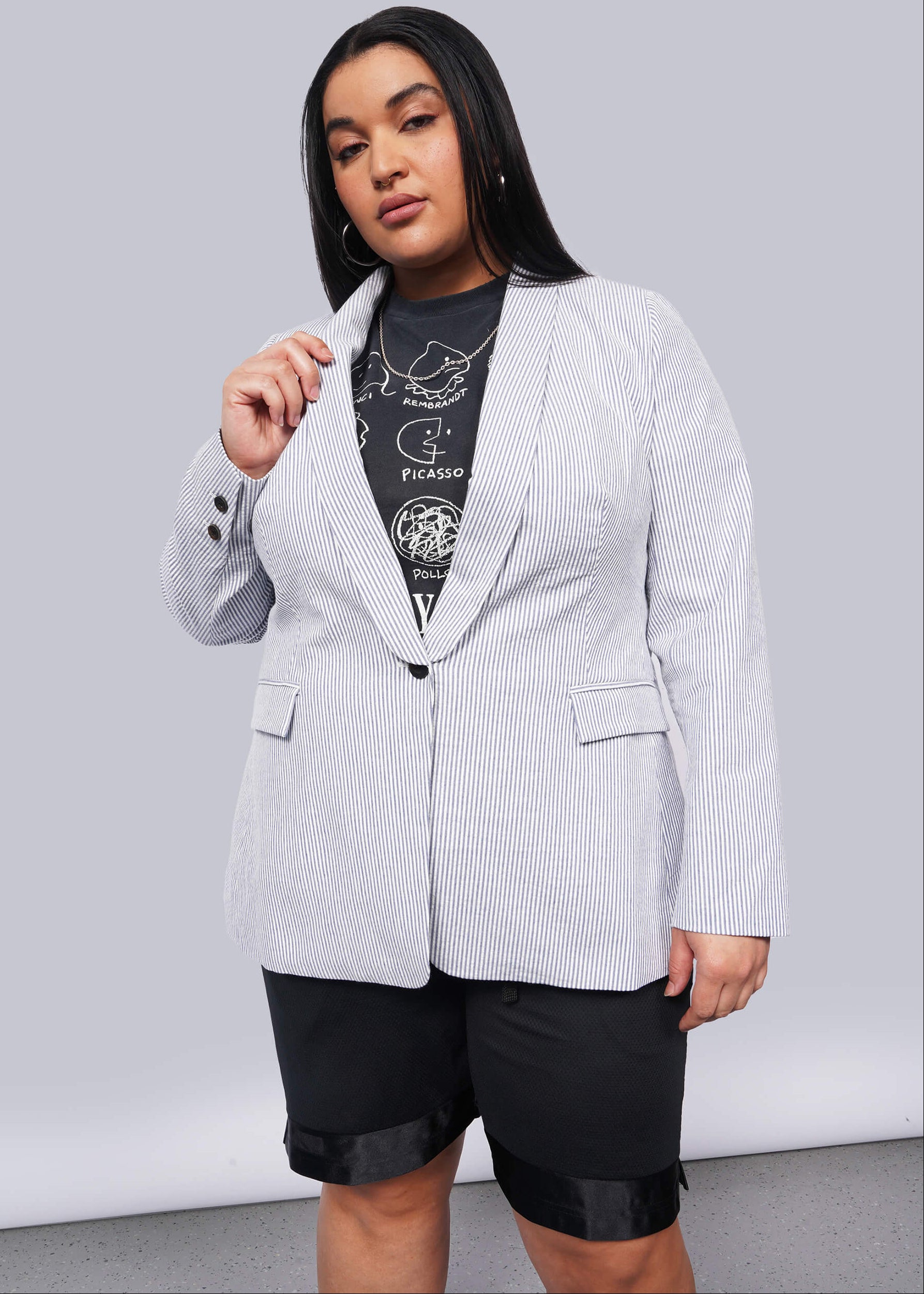 1X=Person wearing a light blue blazer over a graphic t-shirt and black shorts on a plain background