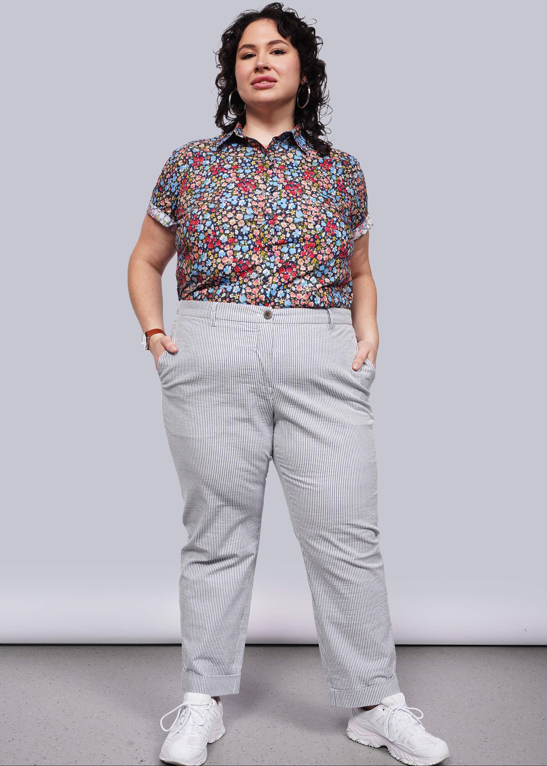 1X=Woman wearing a colorful floral blouse and  light blue seersucker  pants against a plain background