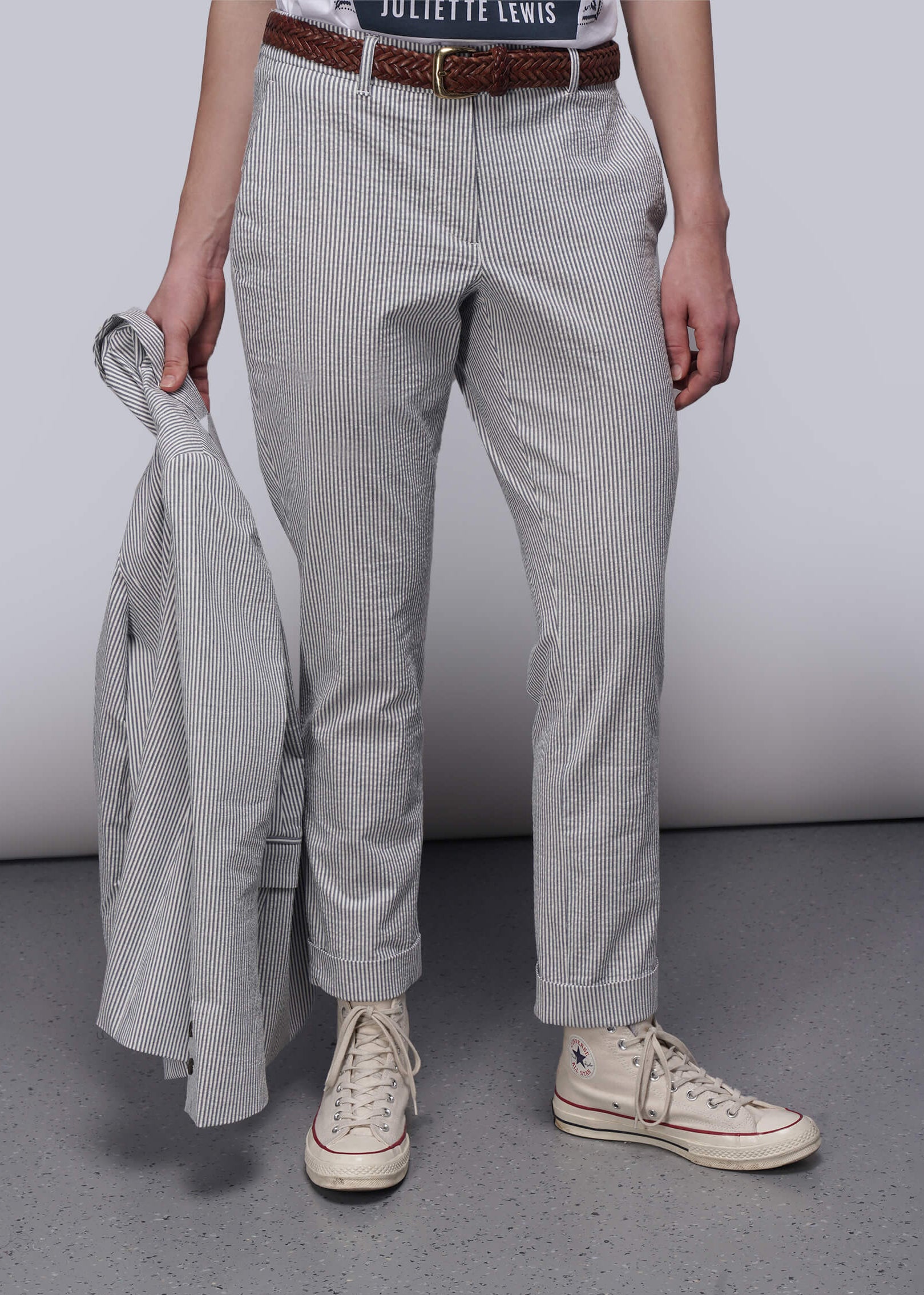 S=Person wearing light blue striped pants and white sneakers holding a gray jacket against a plain background