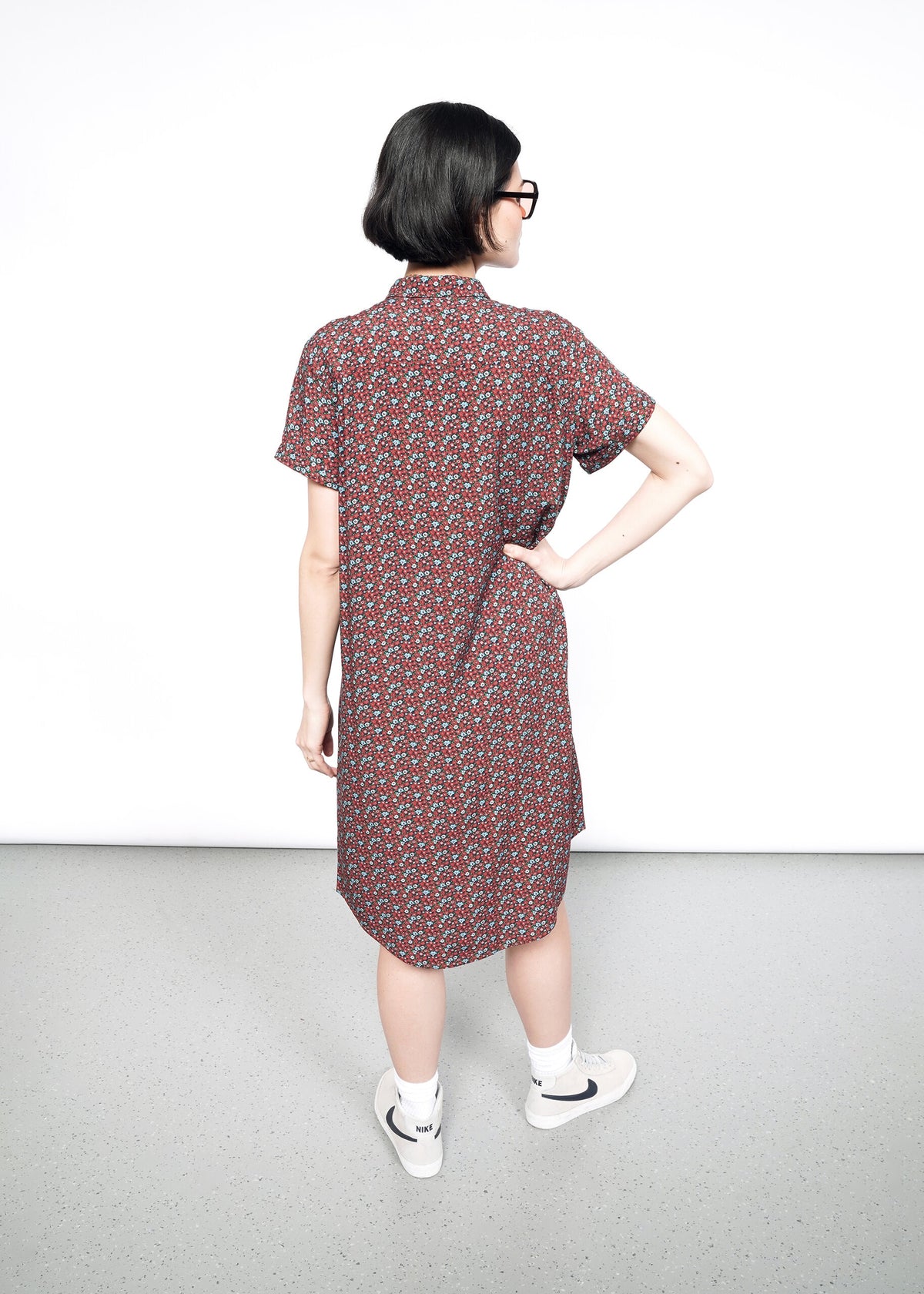 Back image of Model wearing the Empower Short Sleeve Shirt Dress in Floral Cinnamon in size S, with sunglasses, white socks, and tan sneakers