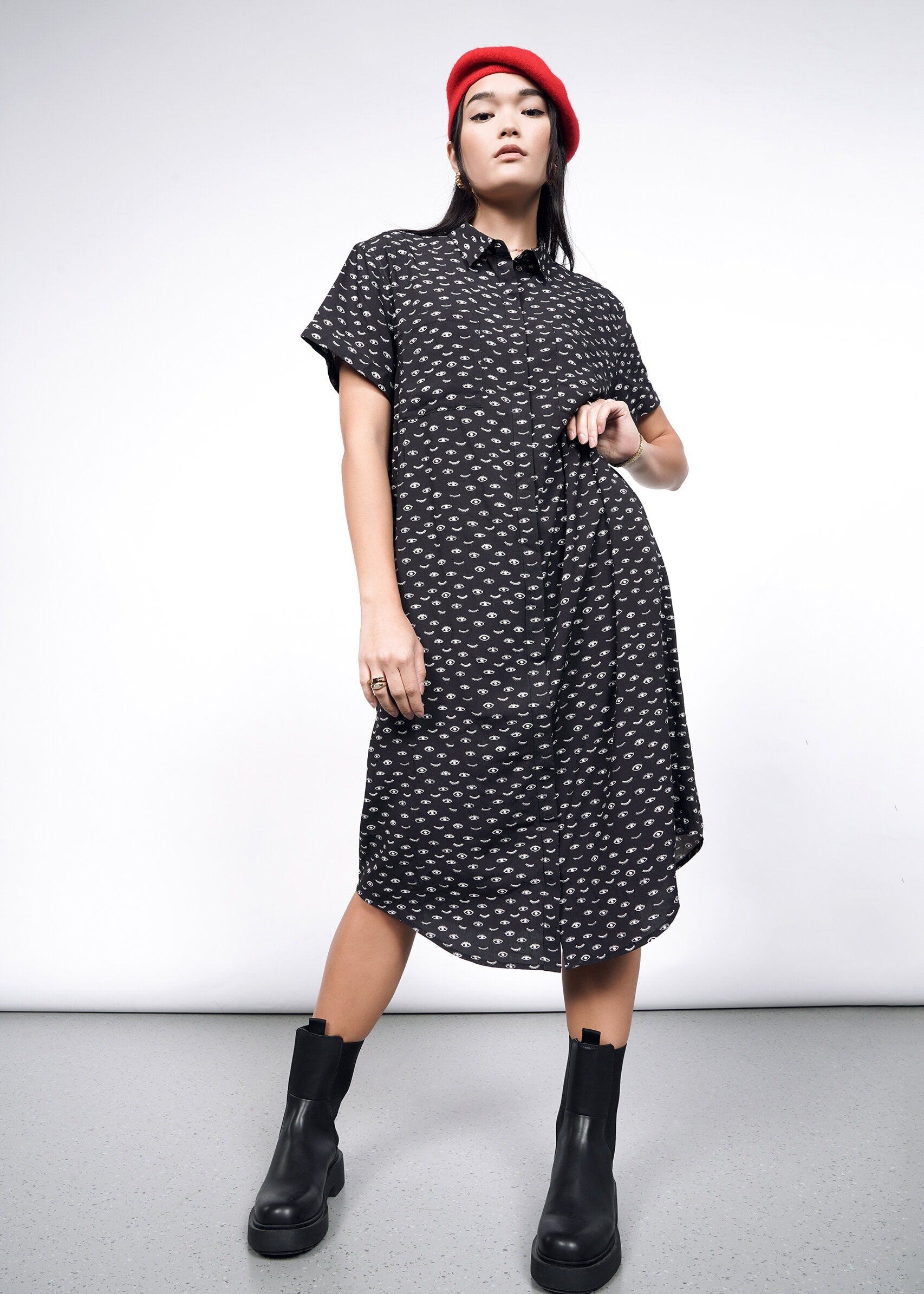 A woman in The Empower Shirt Dress, featuring a black pattern, button closure, and short sleeves below the knees, confidently poses with a red beret and black ankle boots. A plain background highlights her stylish outfit and poised stance.