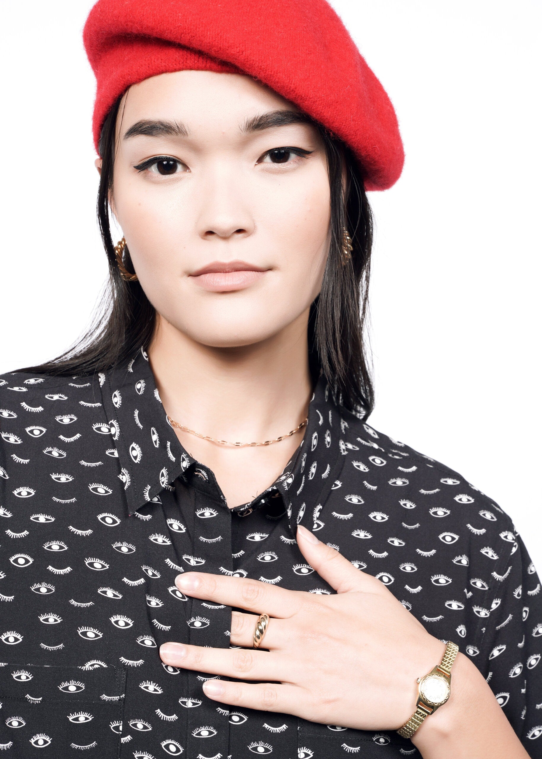 A person with long dark hair wears The Empower Shirt Dress, featuring eye patterns and a chic button closure, along with a red beret. They accessorize with gold hoop earrings, rings, and a watch. Their hand touches their chest against a white background.