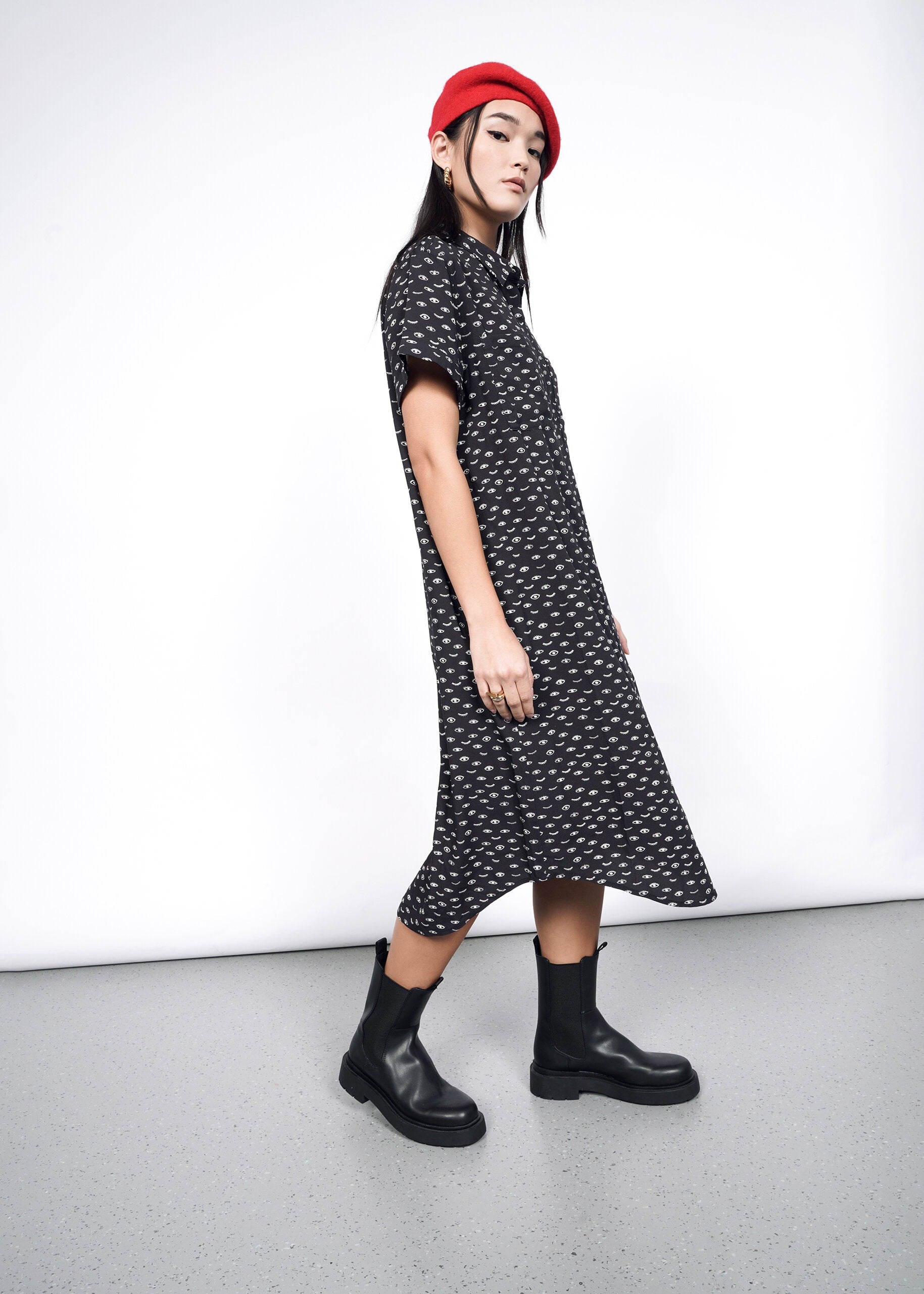 A person wearing a red beret, The Empower Shirt Dress in black with a subtle white pattern, and black boots poses confidently against a neutral background. The stylish dress features a modern button closure, completing the chic look.