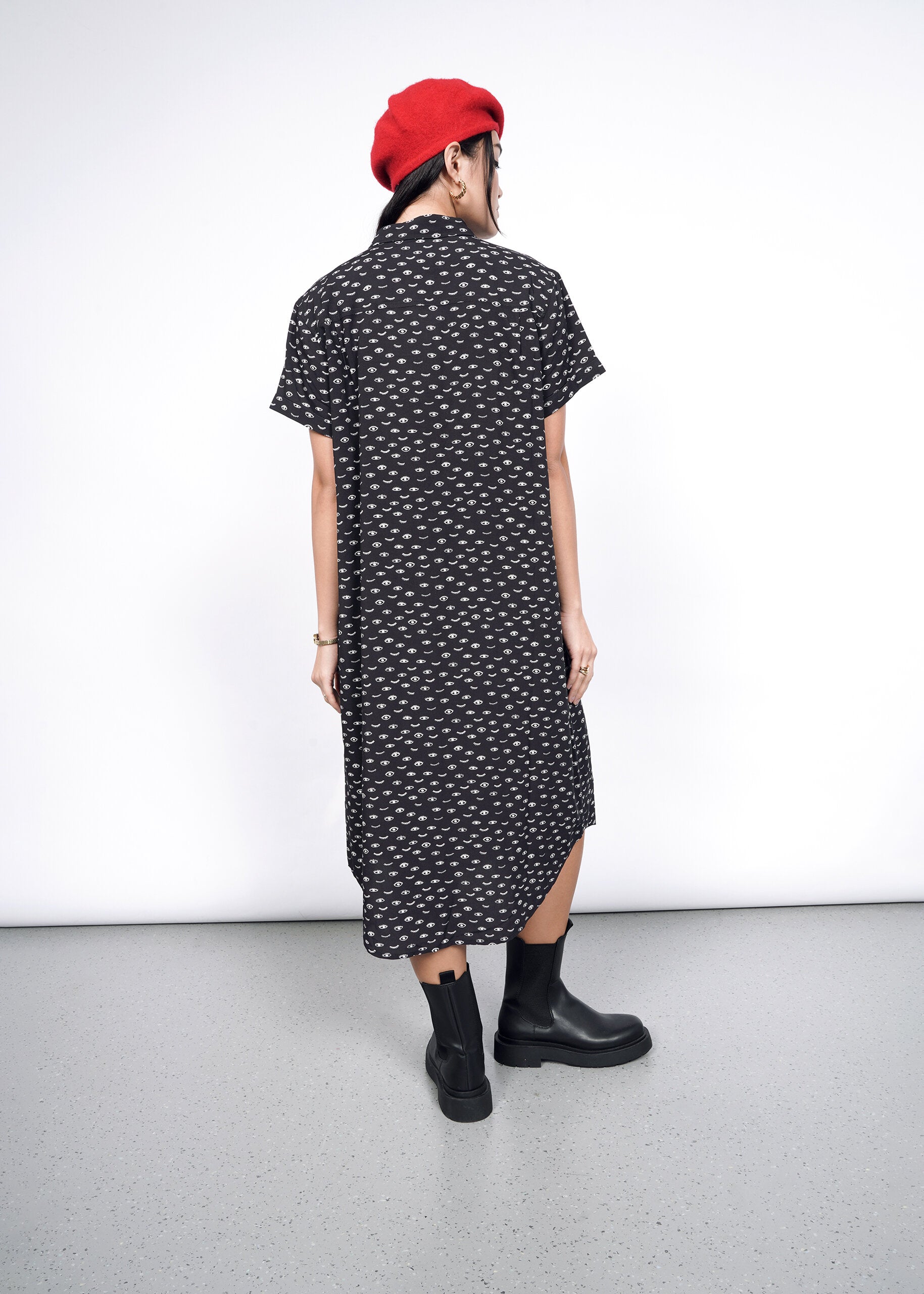 A person wearing The Empower Shirt Dress, with its black and white pattern, is paired with a red beret and black boots while standing against a plain white wall. Their hands rest by their sides on the light gray floor.