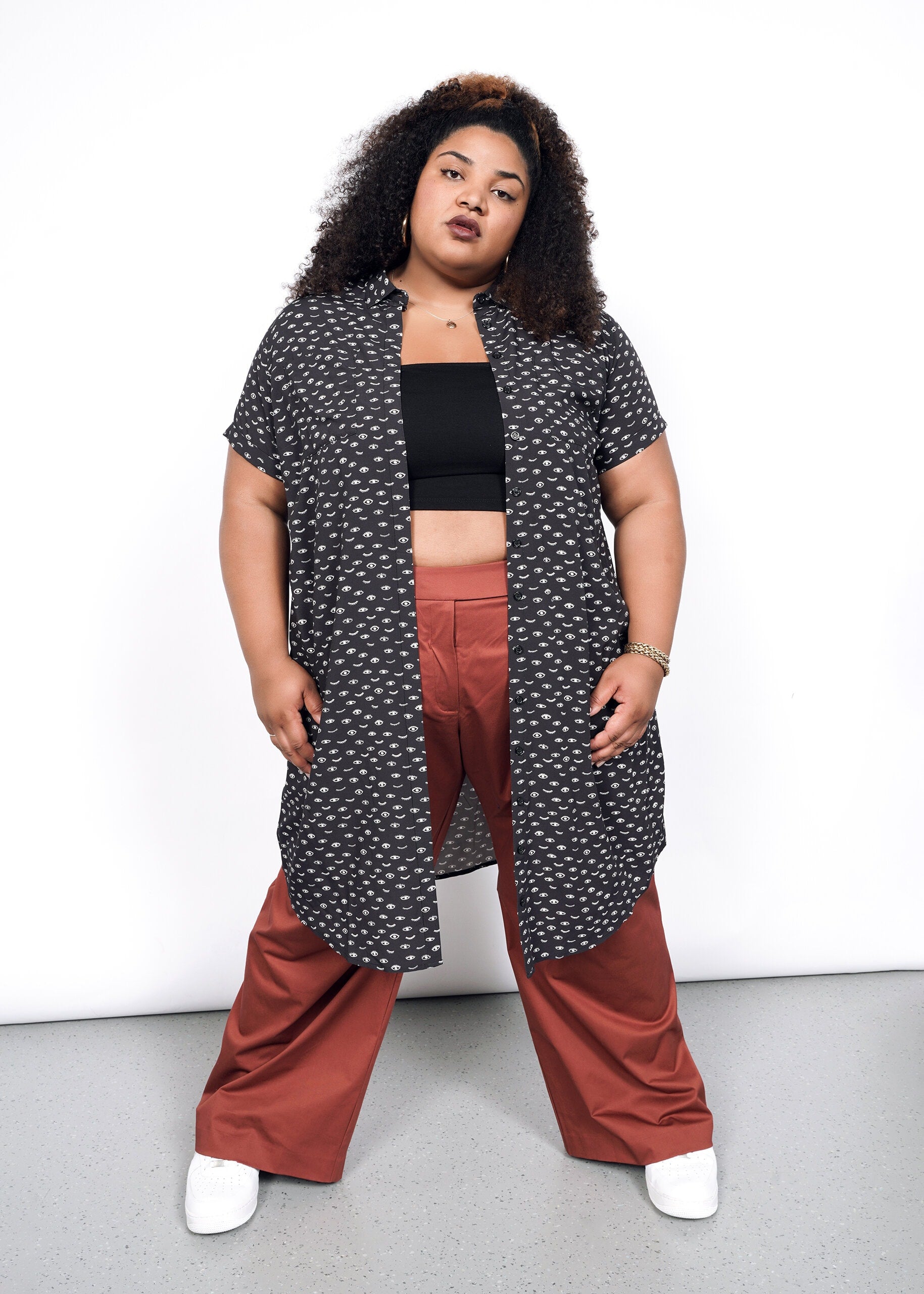 With curly hair, they stand confidently against a white backdrop wearing The Empower Shirt Dress with a black crop top, rust wide-leg pants, and white shoes. Their hands rest on their thighs.