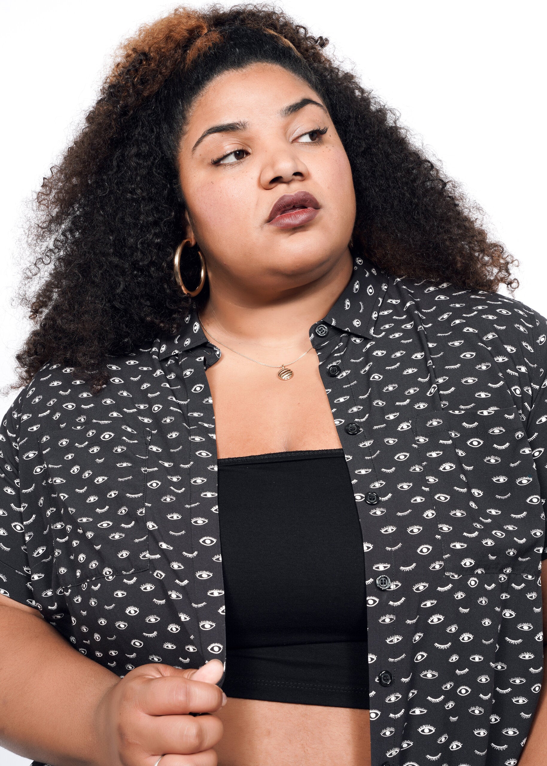 A woman with curly hair models The Empower Shirt Dress over a black top, accessorized with hoop earrings and a necklace, as she gazes to the side against a white background.
