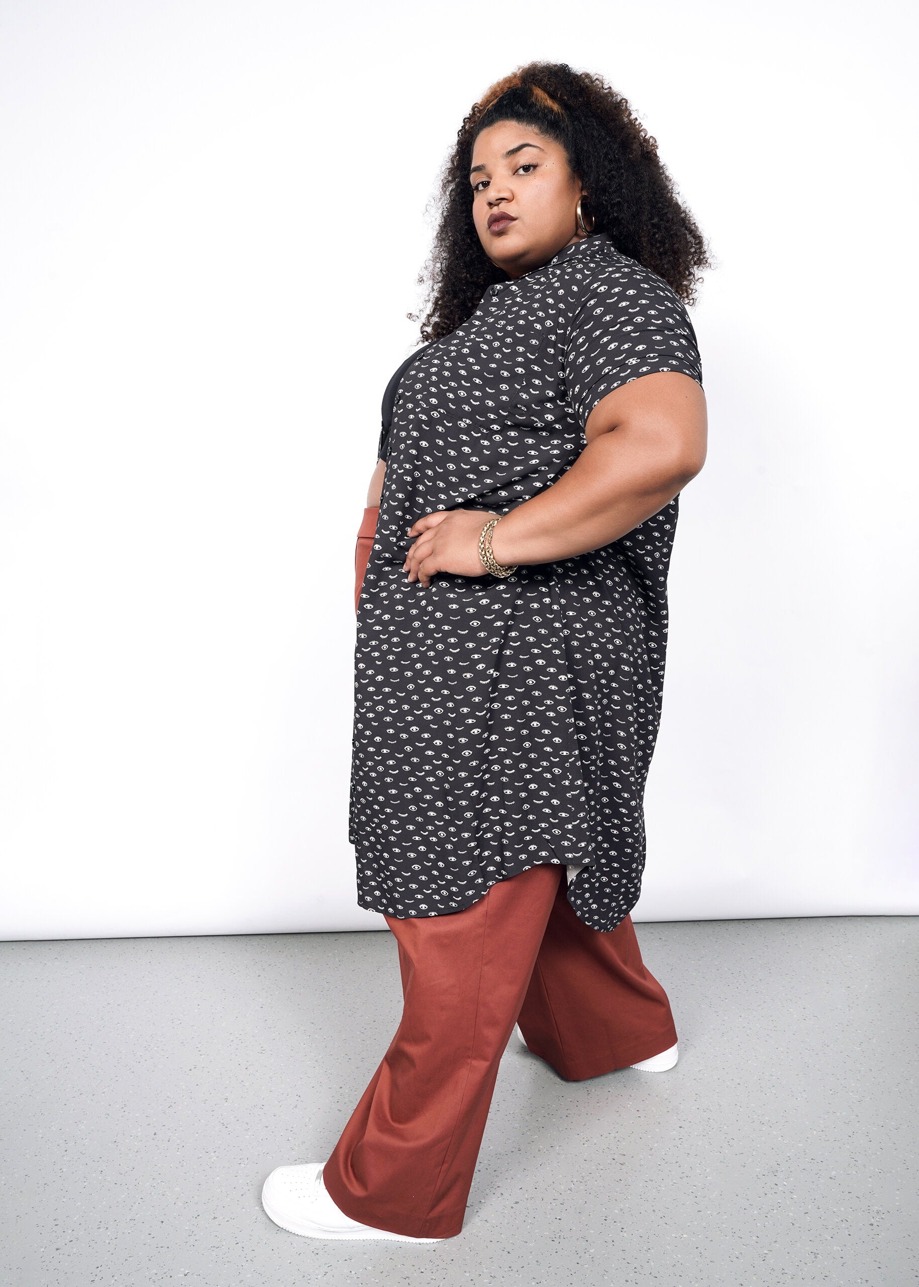 A person with curly hair poses confidently in a stylish tunic resembling The Empower Shirt Dress, paired with rust-colored wide-leg pants. They wear white shoes and a bracelet, standing against a plain white backdrop.