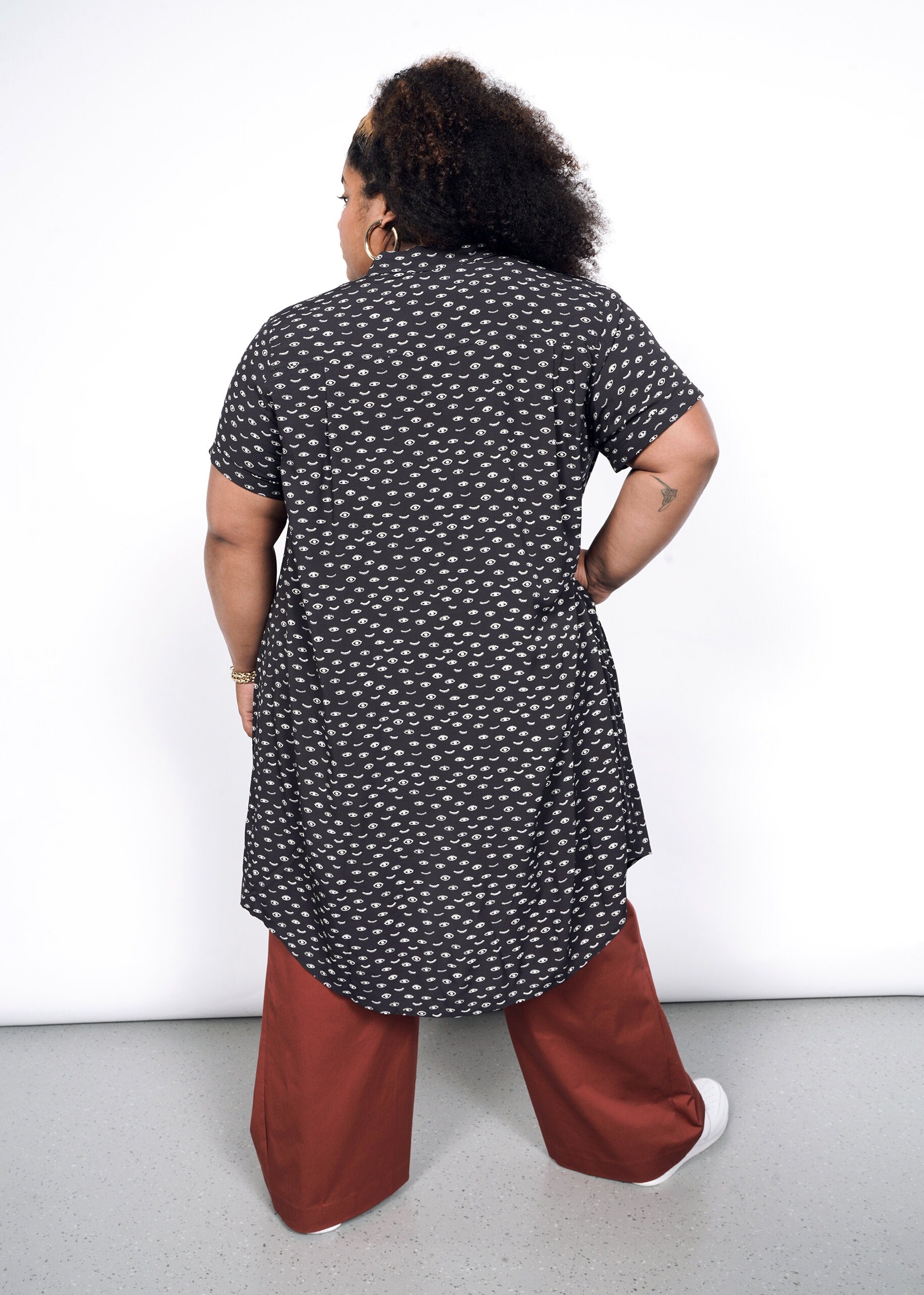 A person with curly hair faces away, highlighting The Empower Shirt Dress in a black and white pattern with button closure, paired with wide-legged rust-colored pants against a plain white background.