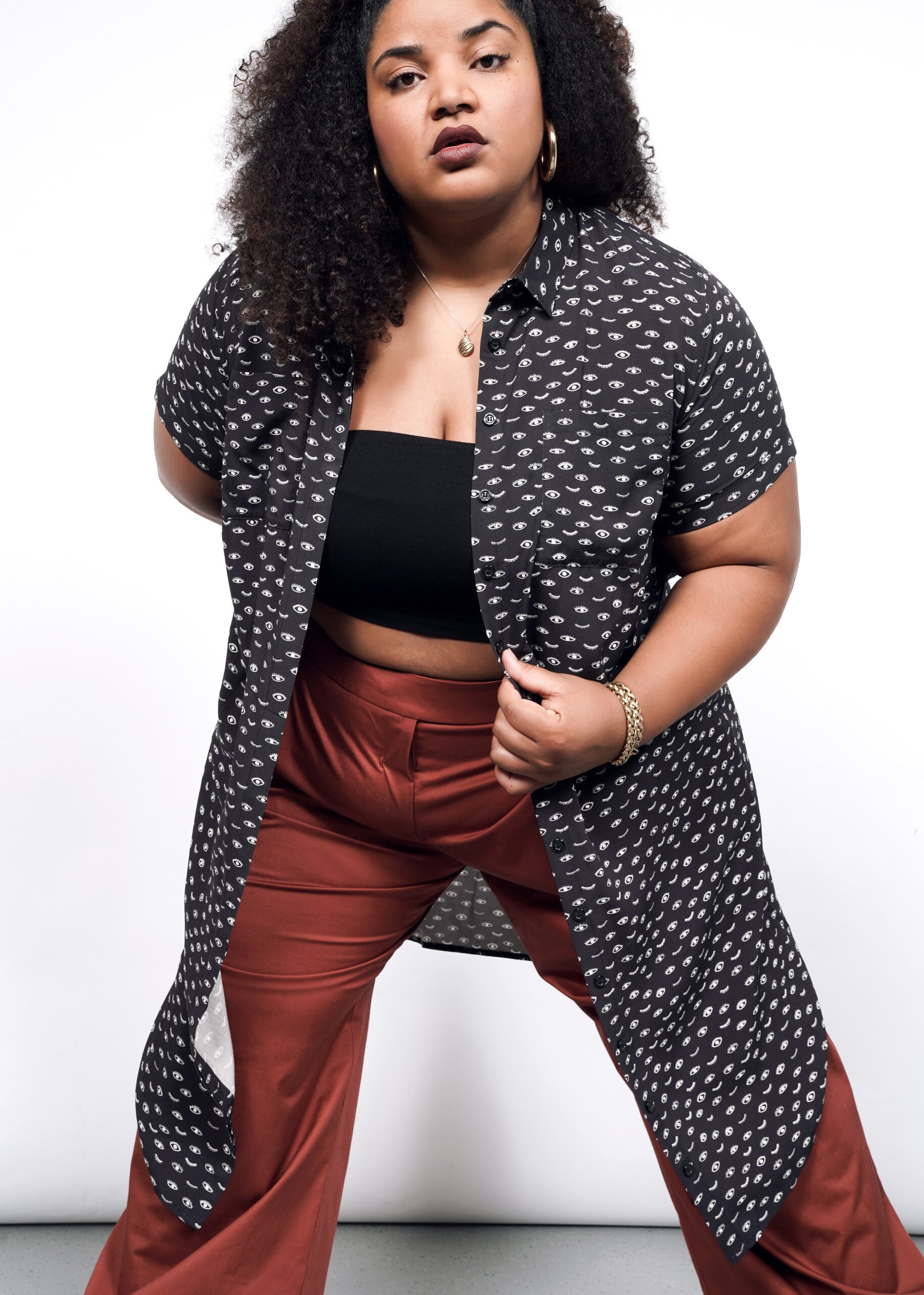 A person confidently poses against a white backdrop, wearing The Empower Shirt Dress with a black crop top and rust-colored pants. Curly-haired, they accessorize with hoop earrings and bracelets.
