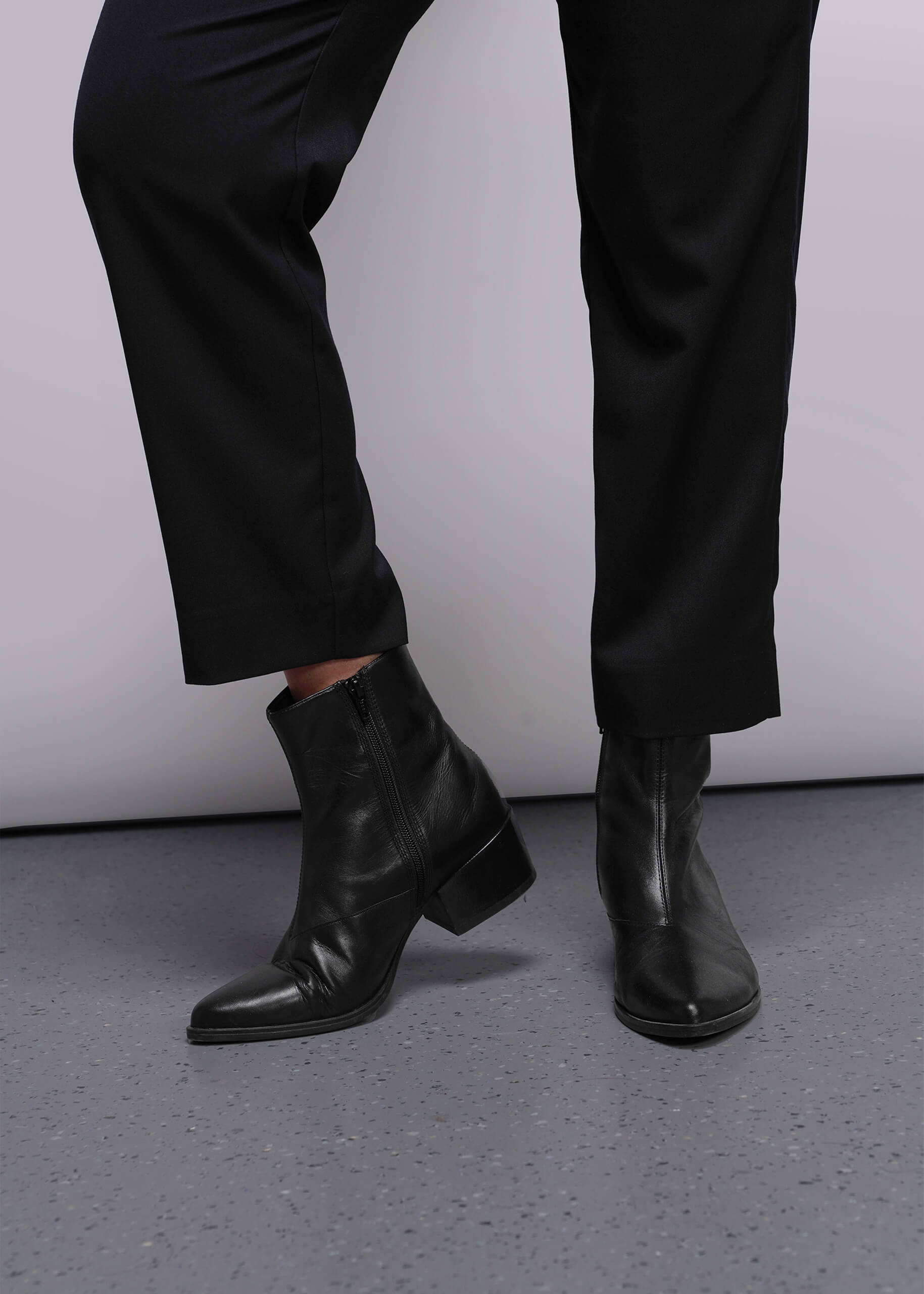 S=Black leather ankle boots worn with black pants on a gray floor.