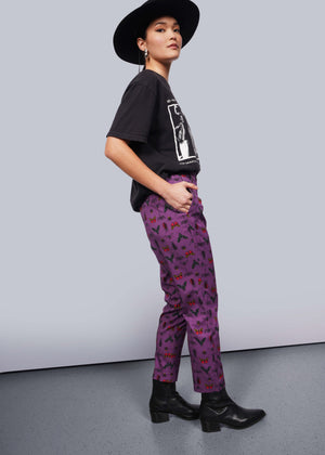 S=Side view of person wearing black wide brimmed hat, black band t-shirt and black heeled boots with hand in pocket of purple bug printed slim crop pants.
