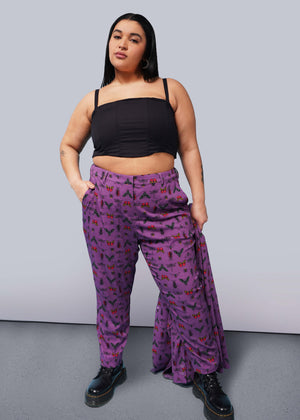 1X= Person with long black hair looks at camera with hand in pocket wearing black corset, purple bug printed slim crop pants, and Doc Marten boots.