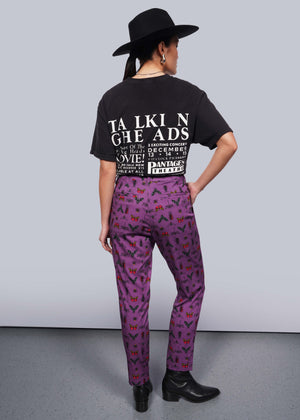 S=Back view of person wearing black wide brimmed hat, black band t-shirt and black heeled boots with hand in pocket of purple bug printed slim crop pants.
