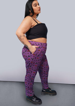1X=Side view of person with long black hair  with hand in pocket wearing black corset, purple bug printed slim crop pants, and Doc Marten boots.